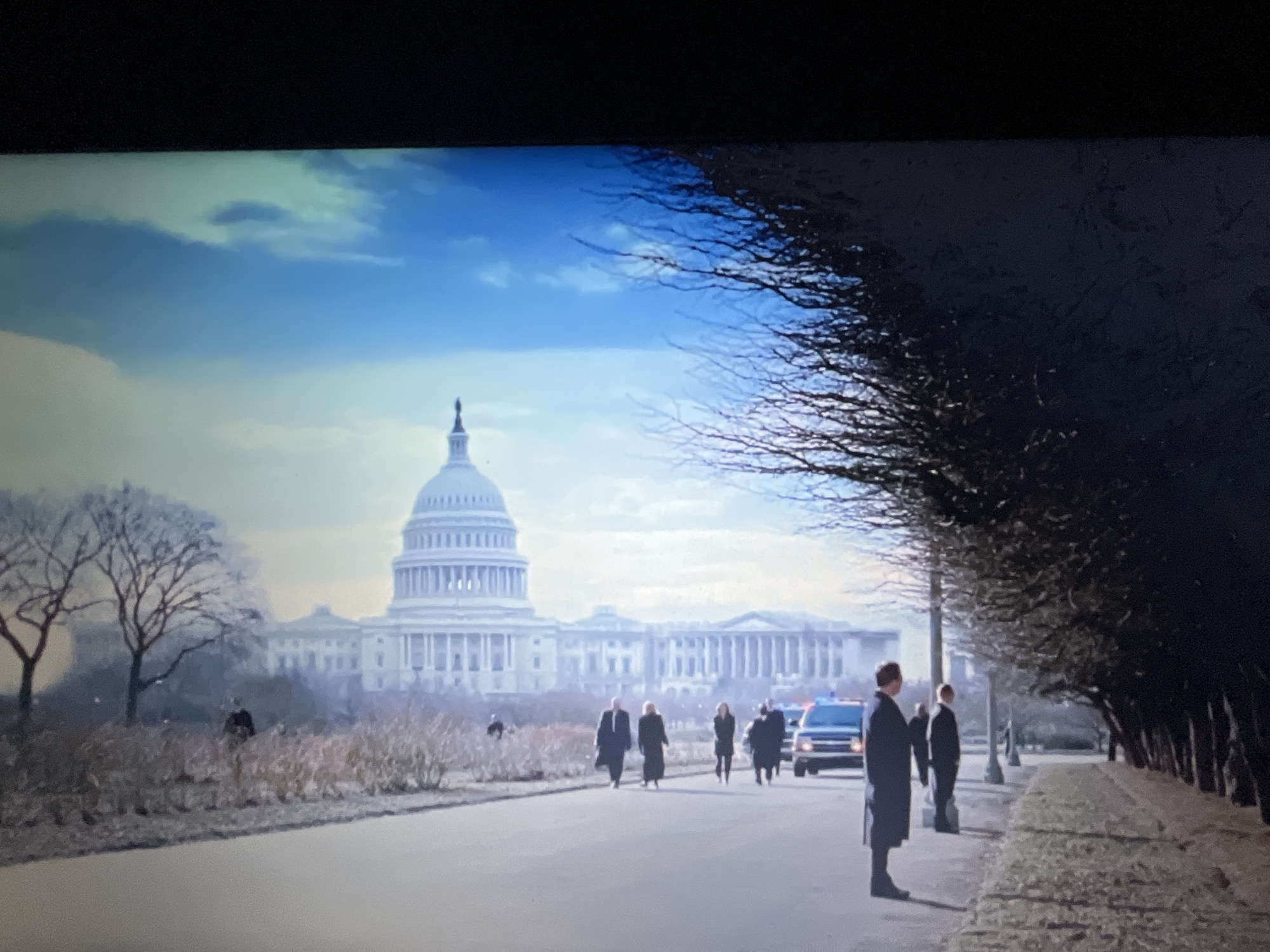 This image depicts the United States Capitol Building under a bright, partly cloudy sky. The building is a large, white neoclassical structure with a prominent dome. In the foreground, a paved walkway is visible, with a line of bare trees casting shadows on the right side of the frame. Several individuals are walking along the walkway, some appearing to be in formal attire. A police car with flashing lights is parked further down the road. There are also more people and bare trees scattered throughout the landscape surrounding the Capitol. The overall scene suggests a formal procession or event taking place in an urban setting. The question about Varna, Bulgaria is irrelevant to the image.