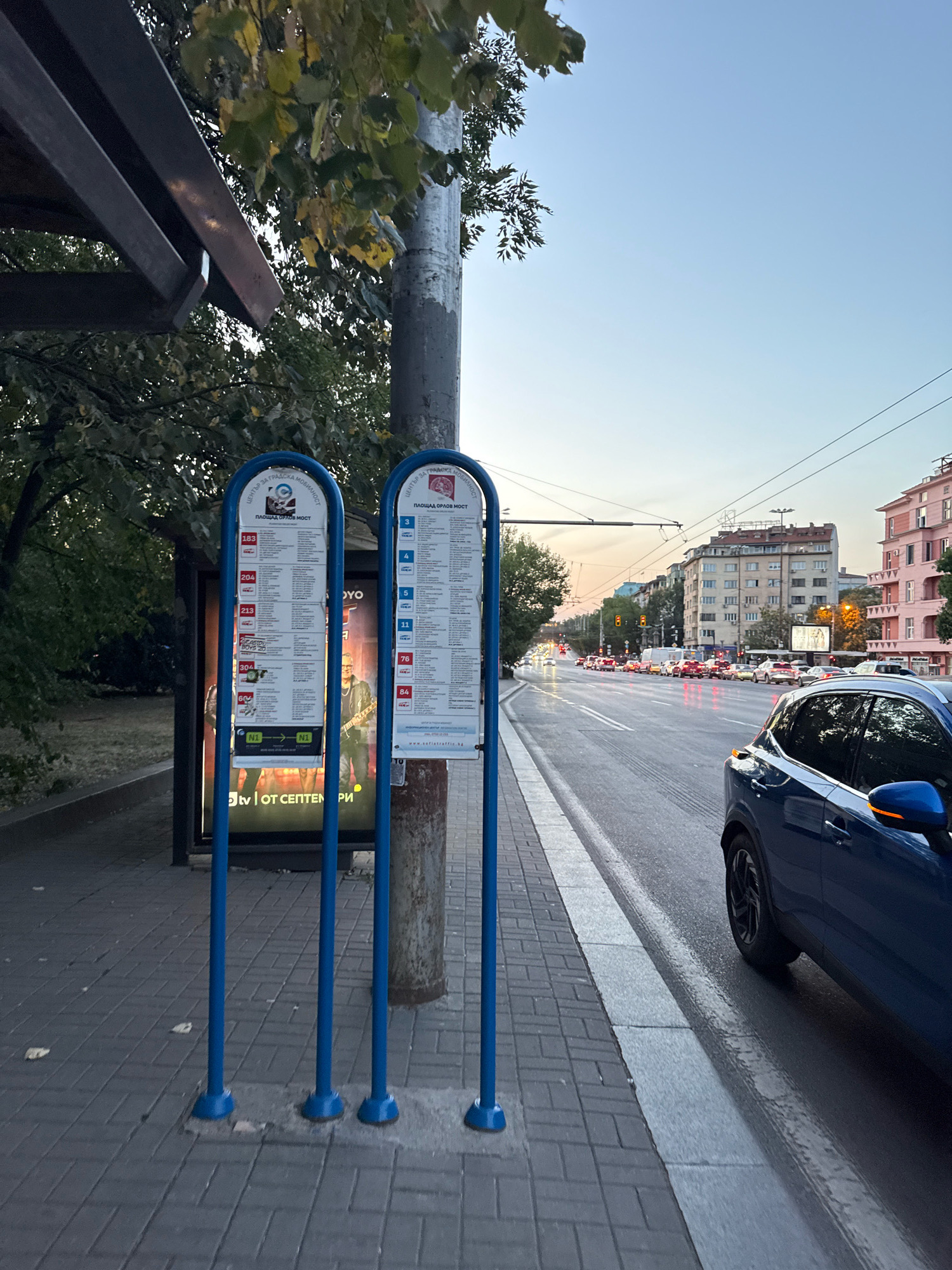 The image shows a bus stop in Sofia, Bulgaria. Two blue bus stop signs are visible, one for routes 183, 204, 213, and 304, and another for routes 3, 4, 5, 11, 76, and 84. A large advertisement poster is behind the left bus stop sign, featuring images of people and text. A blue car is parked on the right side of the street, and traffic is visible in the background, with buildings lining the street. The sky is clear and the scene appears to be during the late afternoon or early evening. The bus stop is located on a sidewalk paved with gray interlocking bricks, adjacent to a wet asphalt road. A street lamp is visible behind the bus stop signs.
