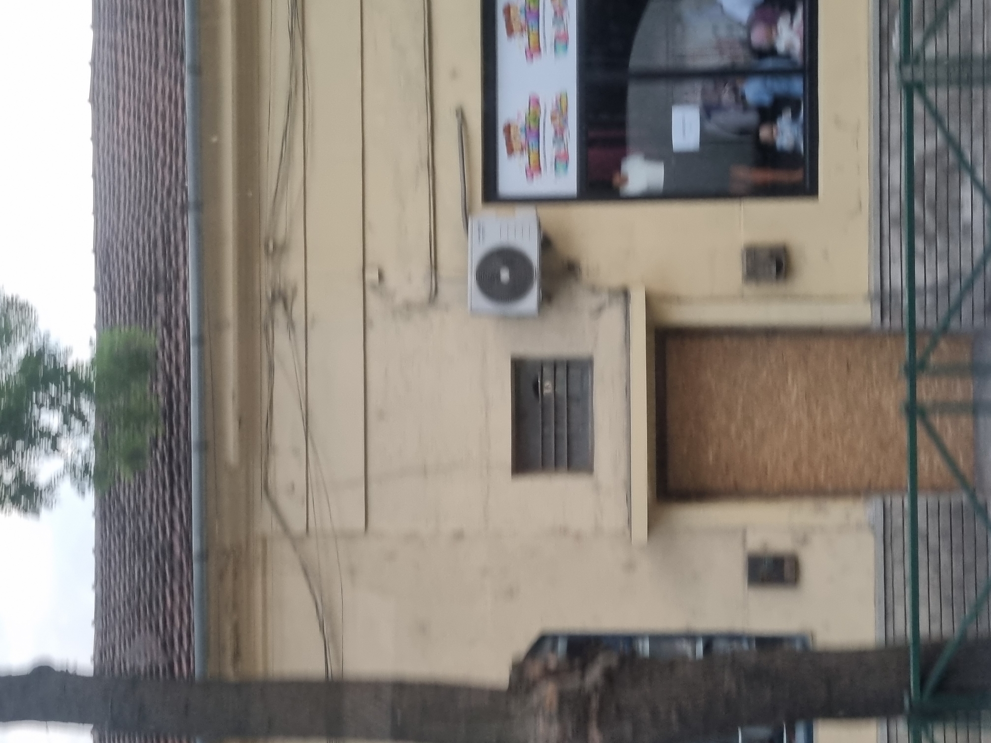 The image shows a yellow building with a tiled roof. A tree trunk is visible on the left side of the image, obscuring part of the building. The building has a boarded-up door in the center. To the right of the boarded-up door, there is a storefront window displaying mannequins dressed in children's clothing. An air conditioning unit is mounted on the wall above the storefront. The building appears to be located in Salonta, Romania, as indicated by the metadata. The overall scene suggests a commercial establishment, possibly a clothing store, with some areas appearing to be under renovation or temporarily closed.