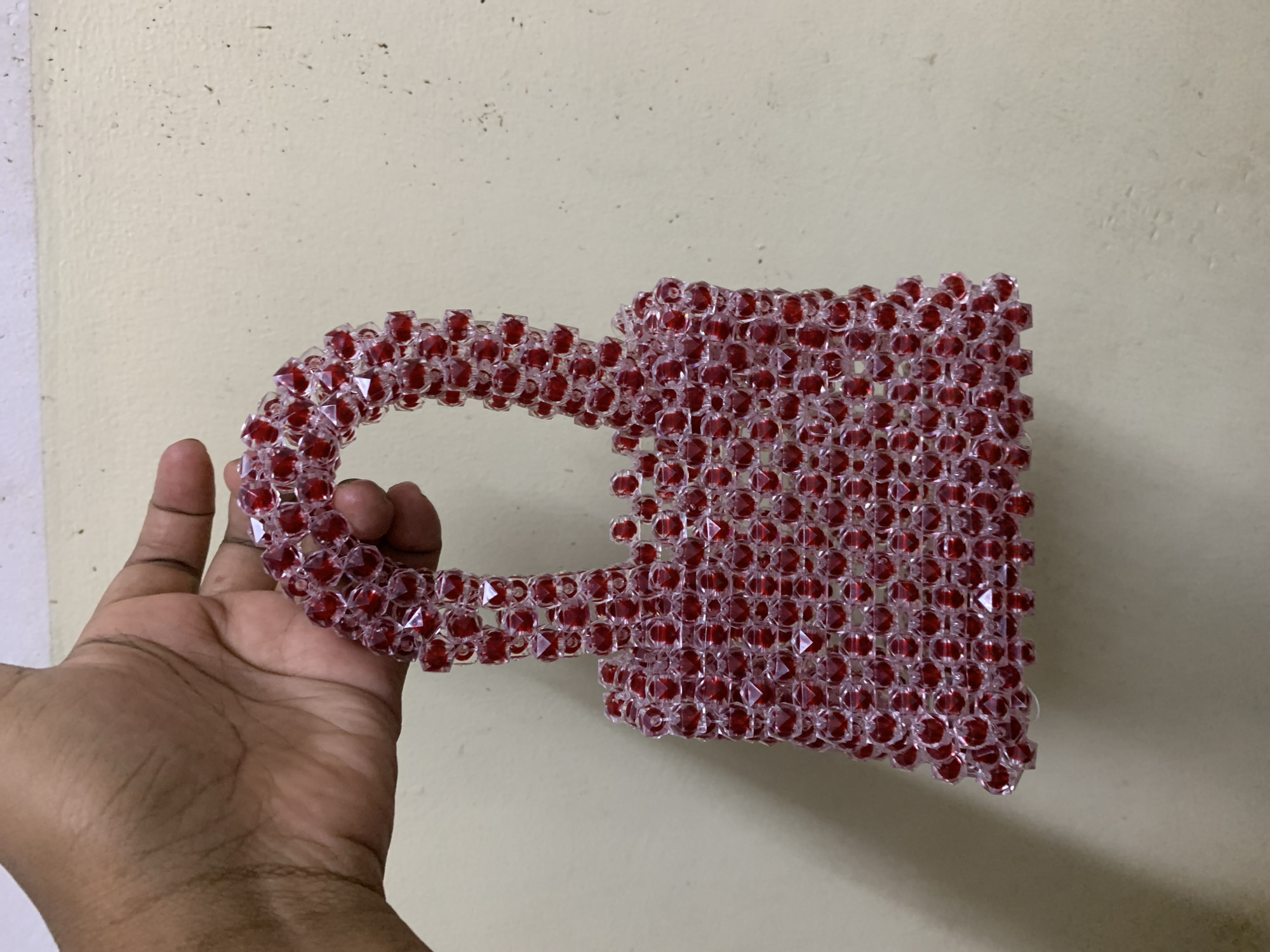 A close-up, eye-level shot captures a person's hand holding a handcrafted item made of red and clear faceted beads against a light-colored wall. The main subject is the intricately beaded object, which appears to be a small bag or pouch with a curved handle on its left side. The beads are small, translucent, and have a distinct red core, creating a sparkling, ruby-like effect as they catch the light. The object's structure is visible, showcasing how the individual beads are strung together to form a solid yet flexible shape.
A dark-skinned hand is positioned on the left side of the frame, supporting the beaded item from below and holding its handle, presenting the craft to the viewer. The background is a plain, textured wall, likely painted in a light beige or off-white hue, showing some minor surface imperfections and dark specks. The lighting is even and soft, suggesting an indoor setting or an area with diffused natural light. There are no other people or discernible objects in the frame, and no visible text. The overall scene is simple, focusing entirely on the craft and the hand holding it. The image context places the scene in Dar es-Salaam, Tanzania.