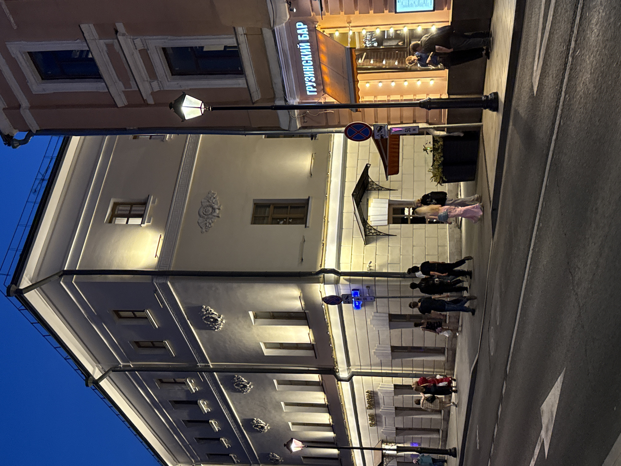 This image captures a street scene in Moscow, Russia, at dusk. A tall, light-colored building with illuminated windows and decorative architectural elements dominates the left side of the frame. To its right, a salmon-colored building features a brightly lit awning with the Cyrillic text "ГРУЗИНСКИЙ БАР" (Georgian Bar). A streetlamp with a hexagonal lantern stands in front of this establishment. Several individuals are visible on the sidewalk and street. On the left sidewalk, a group of people are walking away from the camera. Near the Georgian Bar, two individuals are standing outside, seemingly interacting. The street in the foreground is paved and marked with a white directional arrow. The sky is a deep blue, indicating twilight.