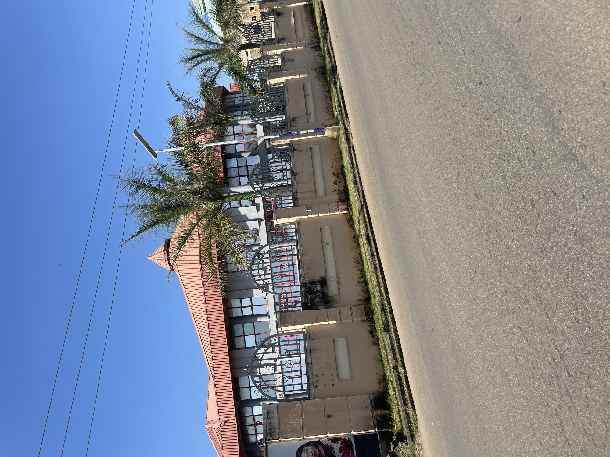 The image shows a building with a red, multi-tiered roof and gray walls, featuring numerous windows with red frames. A decorative metal fence with arched sections runs along the front of the building, behind a light-colored, solid wall. Palm trees are visible in front of the building, and a street lamp is positioned above and to the right of the fence. A large poster of a woman in a red and white floral dress is affixed to the left side of the wall. The scene appears to be on a clear, sunny day, with a bright blue sky. The foreground is dominated by an asphalt road. This scene is consistent with a commercial or institutional building located in Jos, Nigeria.