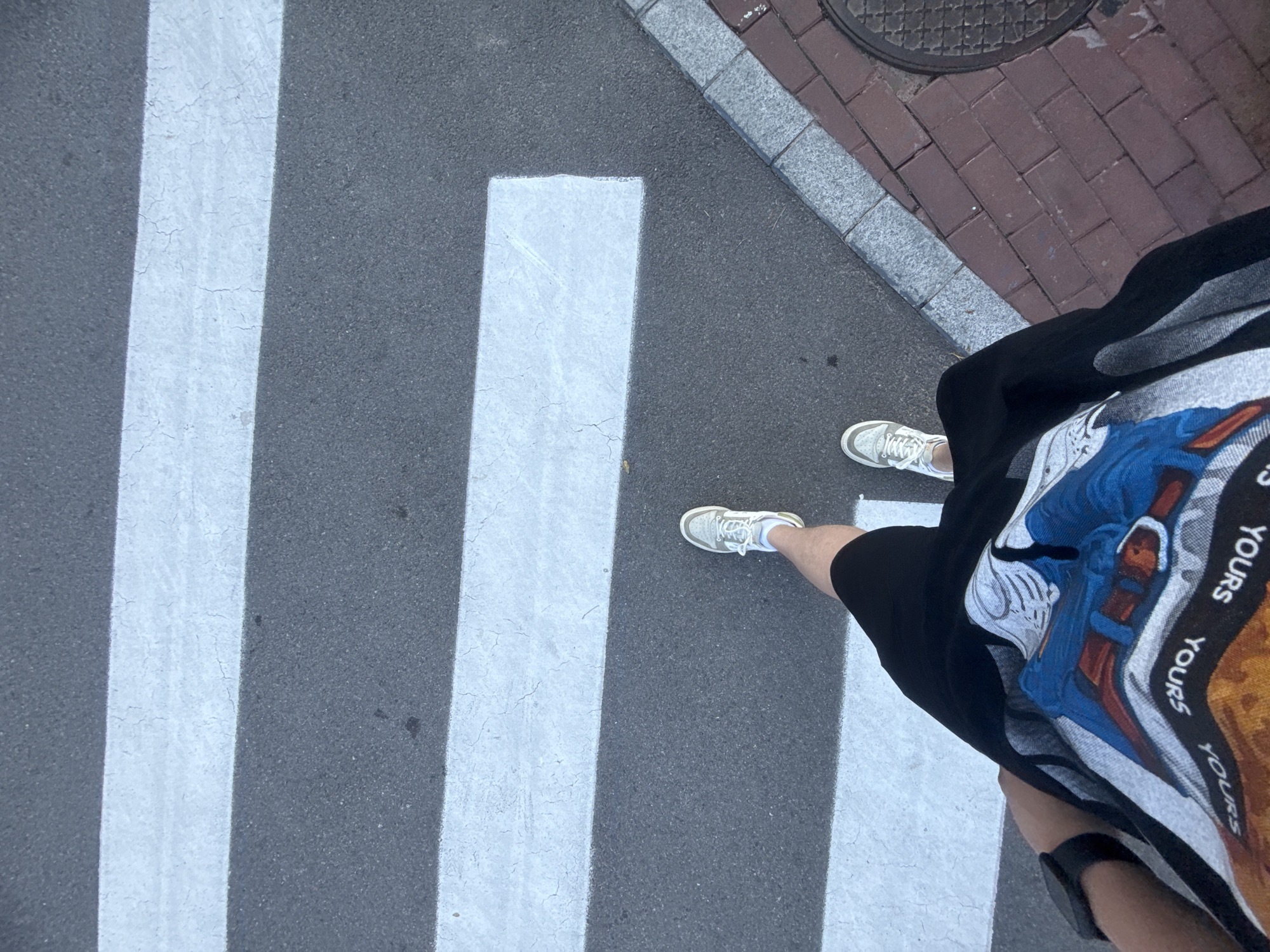The image shows a first-person perspective of a person standing on a pedestrian crossing. The person is wearing white sneakers, black shorts, and a graphic t-shirt. The pedestrian crossing is marked with white stripes on asphalt. To the right of the crossing, there is a sidewalk made of bricks with a grey border, and a manhole cover. The scene appears to be in an urban environment. The lighting suggests it is daytime. The specific location in Qinhuangdao, China cannot be determined from the visual information provided.