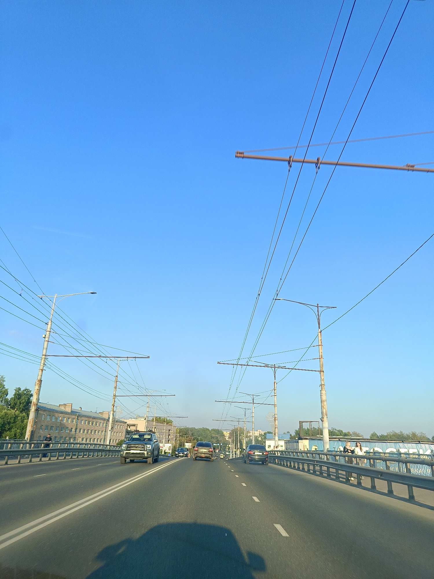 This is a forward-facing, ground-level view of a multi-lane road, likely a bridge, in the city of Vologda, Russia, under a clear blue sky. The road has white lane markings. Several vehicles are visible on the road, including a black SUV in the foreground, a white sedan, and a dark sedan further ahead. On the right side of the road, there is a metal railing with a blue banner behind it, featuring text in Cyrillic. Several people are visible walking along the sidewalk behind the railing. On the left side of the road, there are buildings with multiple windows and trees. Numerous overhead wires, likely for trolleybuses or trams, are suspended by tall poles on both sides of the road. The overall lighting suggests it is daytime, possibly late afternoon or early morning given the angle of the shadows.