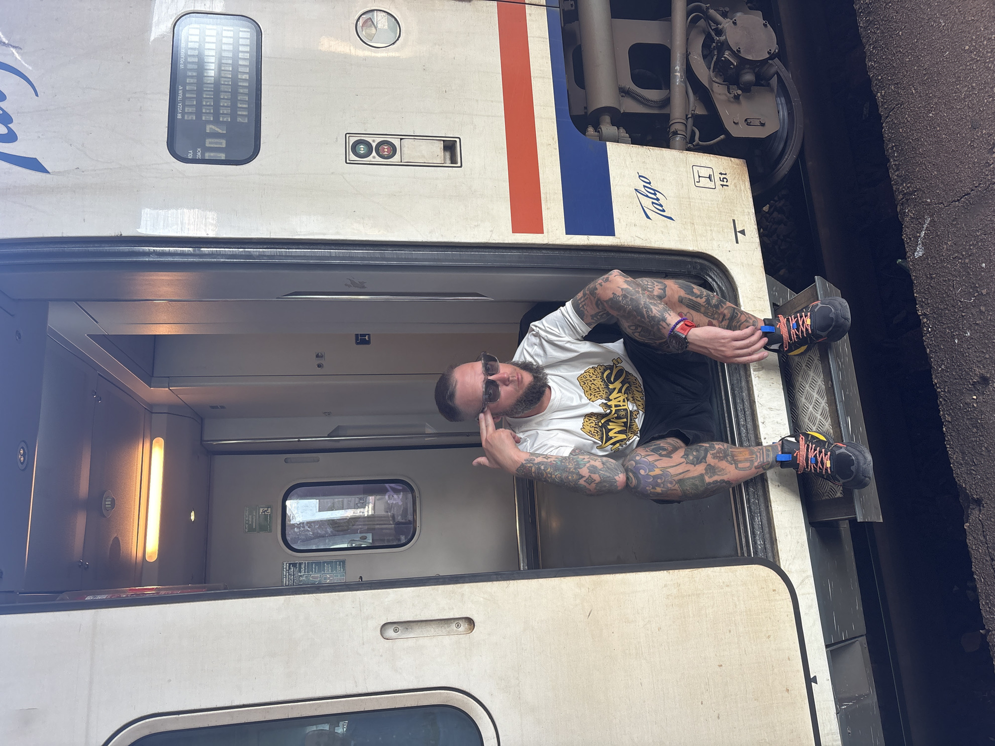 A man is sitting in the open doorway of a white Talgo train car, with the word "Talgo" in blue lettering visible on the exterior. The train car has a red and blue stripe and a number display showing "007." The man has tattoos on his arms and legs, is wearing a white t-shirt with a design, black shorts, and multicolored sneakers. He is wearing sunglasses and pointing his fingers to his head. The interior of the train car is visible, showing a window and a light. The train is next to a dark-colored platform or track. The photograph was taken in Mostar, Bosnia and Herzegovina.