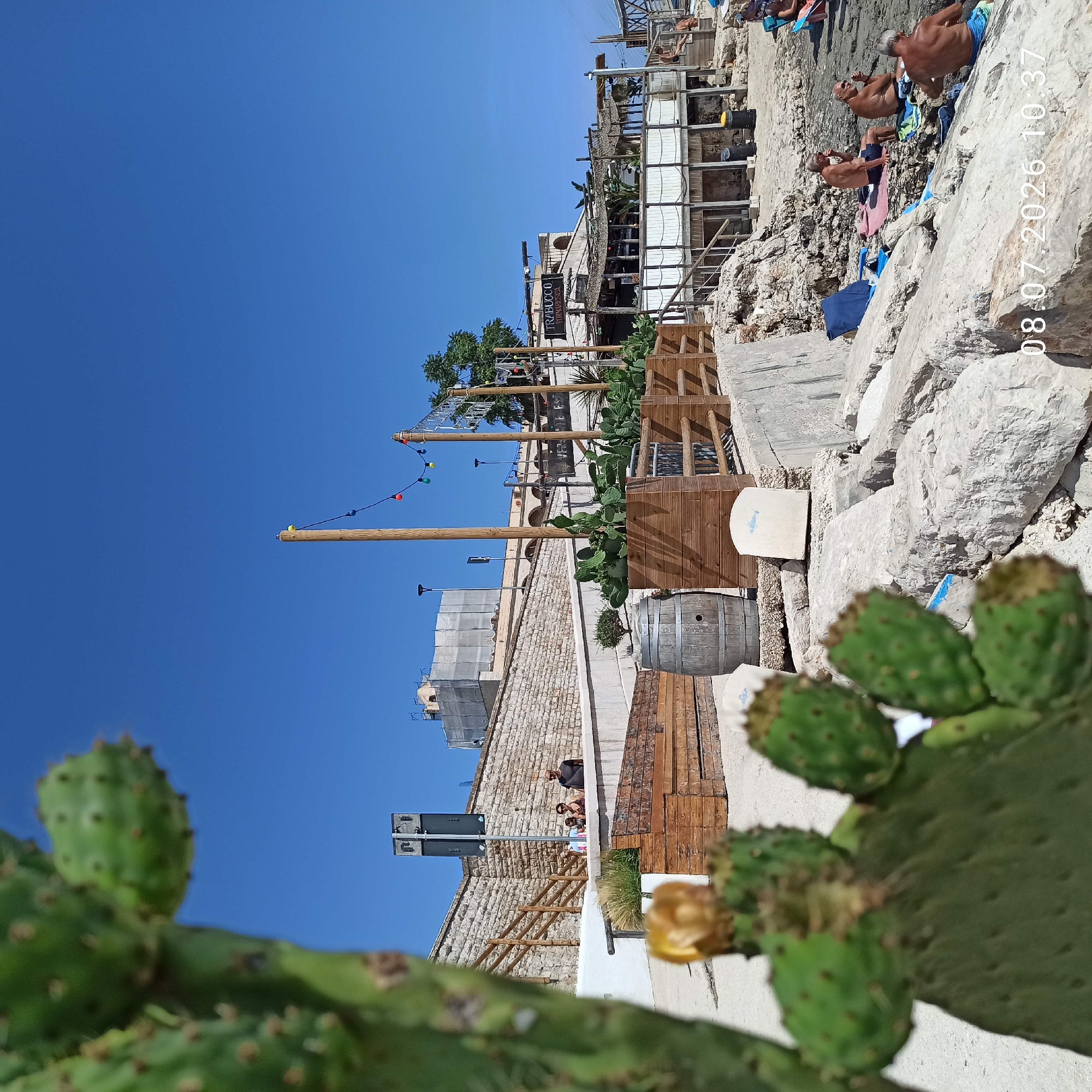 The image depicts a coastal scene in Trani, Italy, likely a beachside establishment. In the foreground, a prickly pear cactus is out of focus, with its green pads and developing fruit visible. Behind it, a wooden boardwalk and a barrel suggest a rustic, beach-themed area. Several people are visible in the mid-ground and background. On the left, three individuals stand near a stone wall. Further back, a multi-story building, possibly a restaurant or club, is situated on the coast. This structure features a sign that reads "TRABUCCO TORMARESCA". The building has a thatched roof and is accessed by wooden walkways. In the distance, construction work is evident, with scaffolding and protective sheeting covering a building. The scene is set against a clear blue sky. Several people are seated on rocks and towels in the lower right portion of the image, appearing to relax on the shore. The overall environment is rocky and arid, with a clear view of the sea. The timestamp "08.07.2026 10:37" is overlaid on the image.