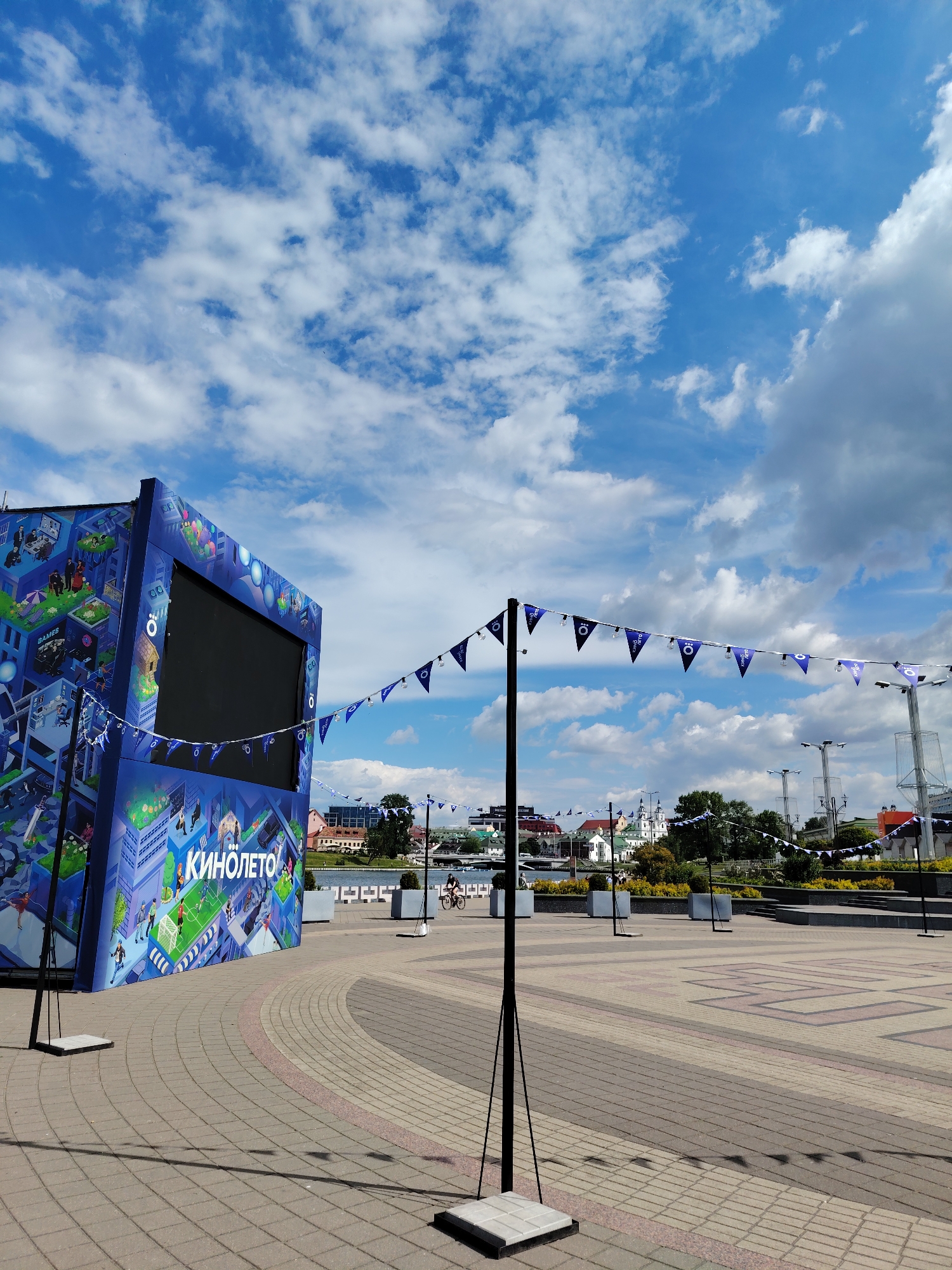 An outdoor plaza or public square in Minsk, Belarus, is depicted under a bright, partly cloudy blue sky. The scene is dominated by a large, box-like blue structure on the left, adorned with colorful, isometric illustrations of a vibrant city showcasing activities like gaming and football. A prominent black screen is set into the front of this structure. White Cyrillic text, "КИНОЛЕТО" (KINOLETO), visible on the front and side, indicates a "Cinema Summer" or "Movie Summer" event.
A string of blue triangular bunting, featuring a white circular logo, stretches across the mid-ground, supported by black poles with square bases. The paving stones of the plaza are a mix of light and dark bricks, forming geometric patterns, with distinct shadows cast by the poles and bunting, suggesting strong daylight.
In the mid-distance, a person rides a bicycle across the plaza. Beyond, a body of water separates the foreground from the distant cityscape. Across the water, several buildings are visible, including a notable white structure with green or blue domes, characteristic of historical architecture. Further in the background, trees and some industrial-looking towers can be seen. The weather is sunny with scattered white clouds, creating a pleasant atmosphere for the outdoor event suggested by the installation.
