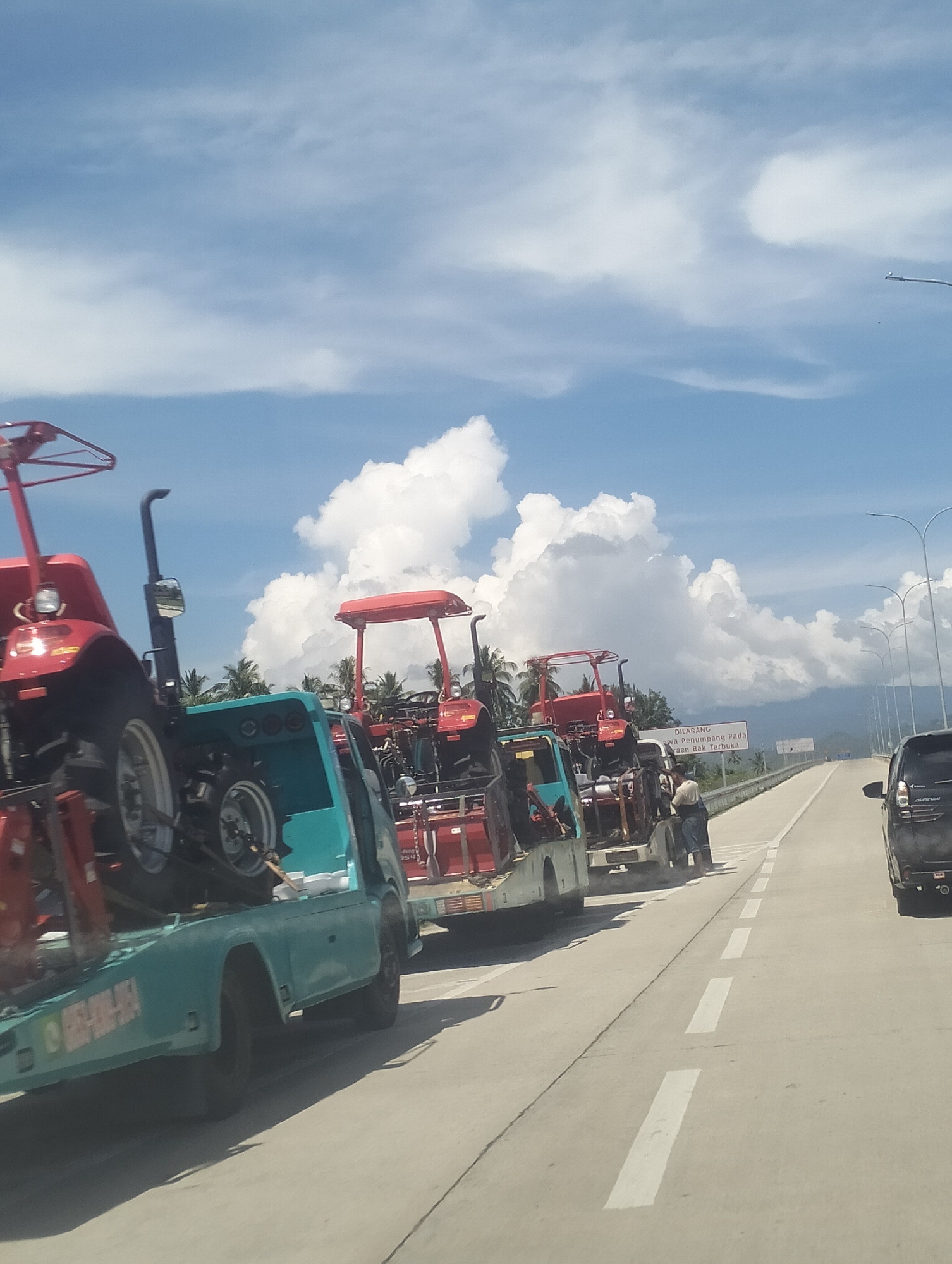 In Aron Bunoet, Indonesia, a convoy of flatbed trucks is transporting several red tractors on a highway. The sky is blue with scattered white clouds, and palm trees are visible in the background. The road is wide and paved, with lane markings and streetlights. Two individuals are seen standing near the tractors on the side of the road. A black vehicle is in the right lane of the highway.