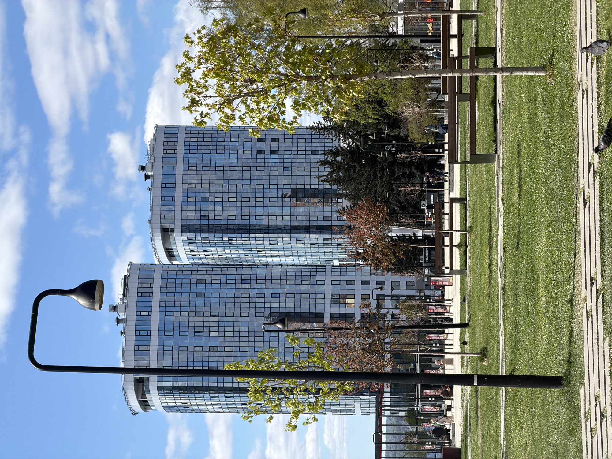 The image captures a clear day in Izhevsk, Russia, with a focus on a modern urban park setting. Two mirrored, cylindrical high-rise buildings with a grid of windows form the backdrop, reflecting the blue sky and scattered clouds. In the foreground, a paved pathway stretches horizontally across the frame, lined with manicured green grass. Black lampposts are visible along the pathway. Several trees, varying in size and foliage color, are interspersed within the park. Benches and some people are seen in the background. Two birds are on the pathway.