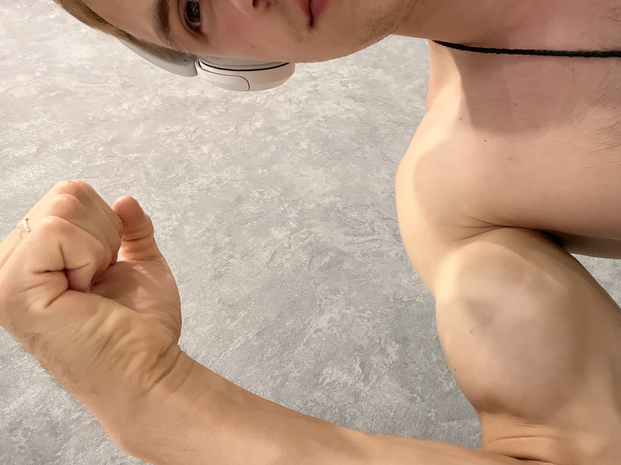 A close-up, upside-down, shoulder-level shot shows a person's arm flexed with their fist clenched. The person is wearing white headphones and a thin black cord around their neck. The background is a gray, textured surface. The lighting appears to be artificial and evenly distributed, with no indication of the time of day or weather. The image is a tight crop, so the setting is indistinct, but the textured gray surface could suggest an indoor environment like a studio or a modern room. The overall mood is neutral, focusing on the physical form and accessory items. There is no visible text in the image.