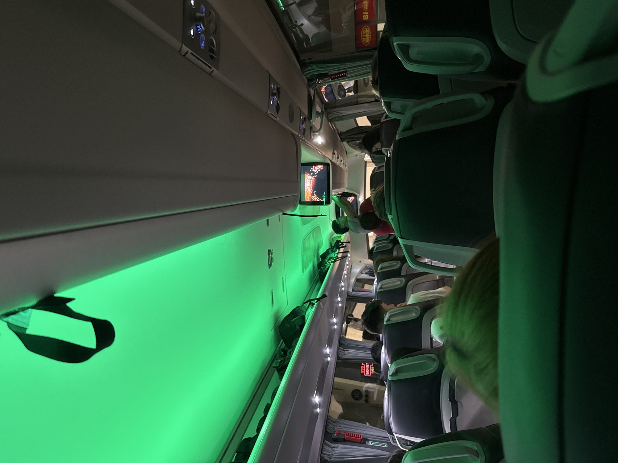 The image depicts the interior of an airplane cabin. Seats are visible on the right, occupied by passengers. Overhead compartments are on the left. A screen is visible on the left. The cabin lighting is green. The location is not discernible from the image.