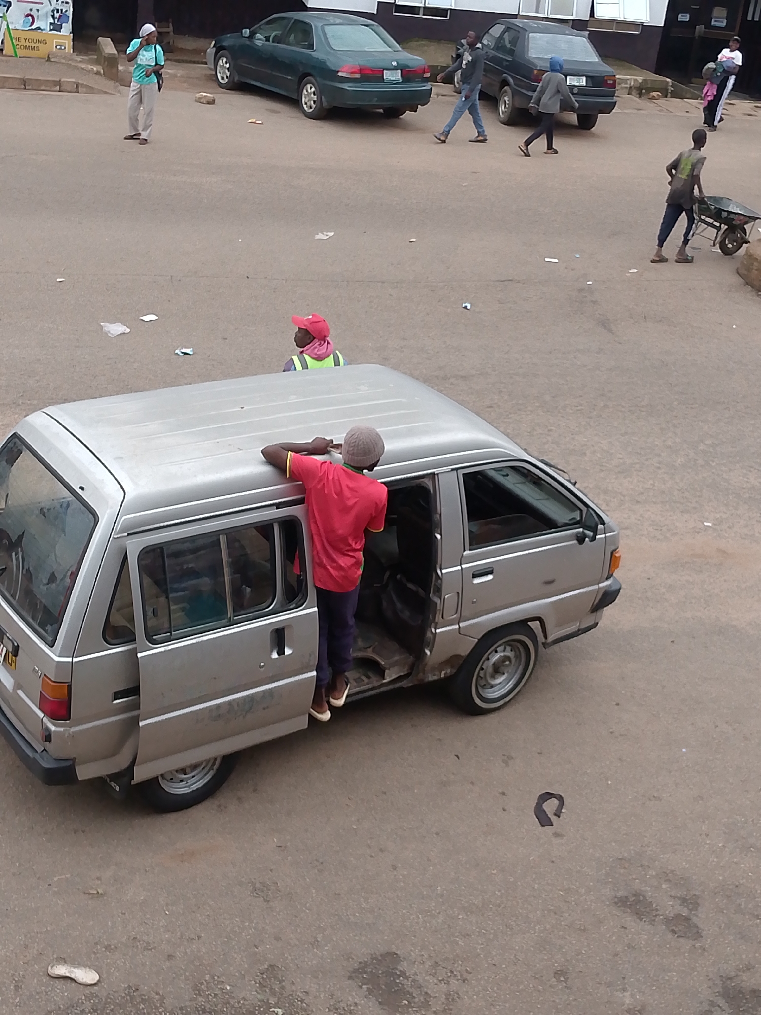 The image depicts a street scene in Bukuru, Nigeria. In the foreground, a silver minivan is parked with its rear passenger door open. A young man wearing a red t-shirt and dark trousers, with a grey cap, is standing on the side of the van, leaning his upper body into the open door. He appears to be reaching into the van. Another person, wearing a pink hat and a high-visibility vest, is visible further back near the van.
In the background, several other individuals are present. A man and a woman are walking near a dark sedan. Further back, a young boy is pushing a wheelbarrow. Another person, wearing a grey hooded jacket and dark pants, is walking away from the camera. In the upper left of the image, a person in a light blue shirt and light-colored pants is standing near a building. The ground is paved with asphalt, and there are scattered pieces of debris on the road. The overall scene suggests a public area, possibly a marketplace or a street with ongoing activity.