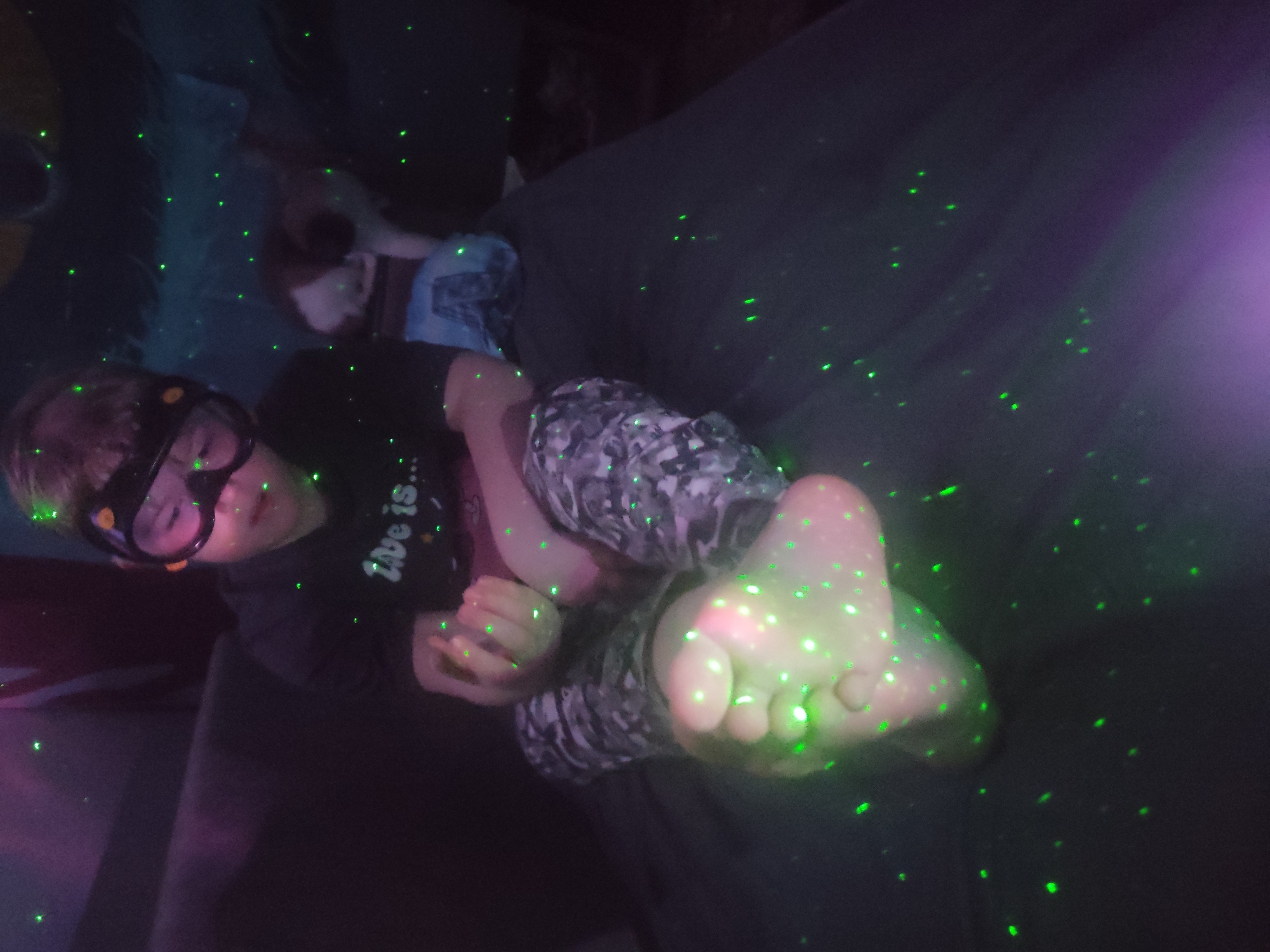 A child wearing goggles and a black t-shirt with the partial phrase "love is..." is lying on what appears to be a dark gray couch or cushion. The child's bare feet are prominent in the foreground, and the child is holding their hands together. The lighting in the scene is dark, with many small, green laser-like dots scattered throughout. A possible location in Chaikovsky, Russia is not discernable from the image.