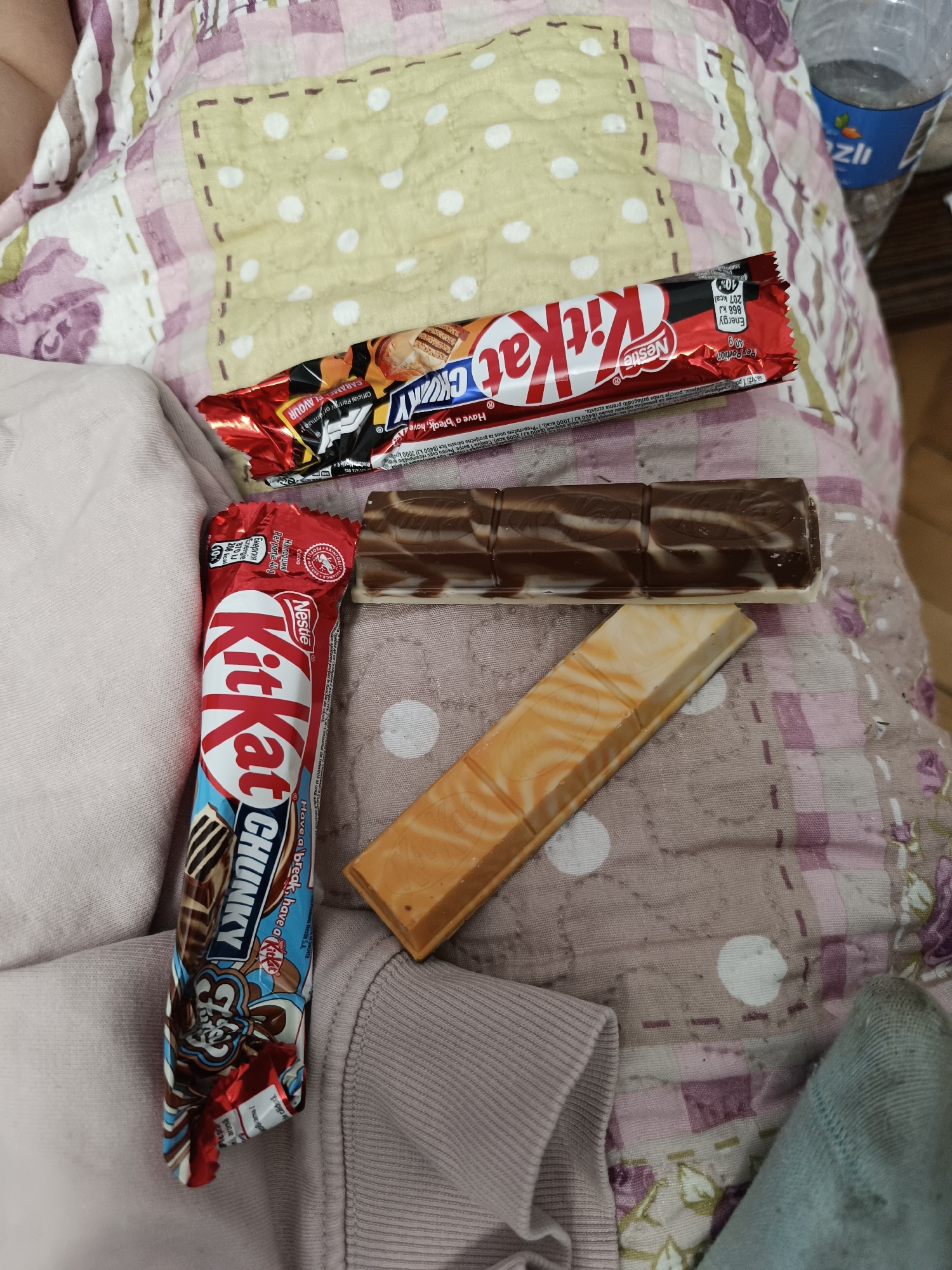 The image shows a person's lap with several candy bars on top of a patterned quilt. There is a KitKat Chunky, a regular KitKat bar, and two other wafer-based candy bars with different flavored fillings. The person is wearing a pink sweatshirt and a blue sock. A water bottle is partially visible in the upper right corner. The image was taken indoors, and the lighting is bright. The location, Pleven, Bulgaria, is not evident in the image.