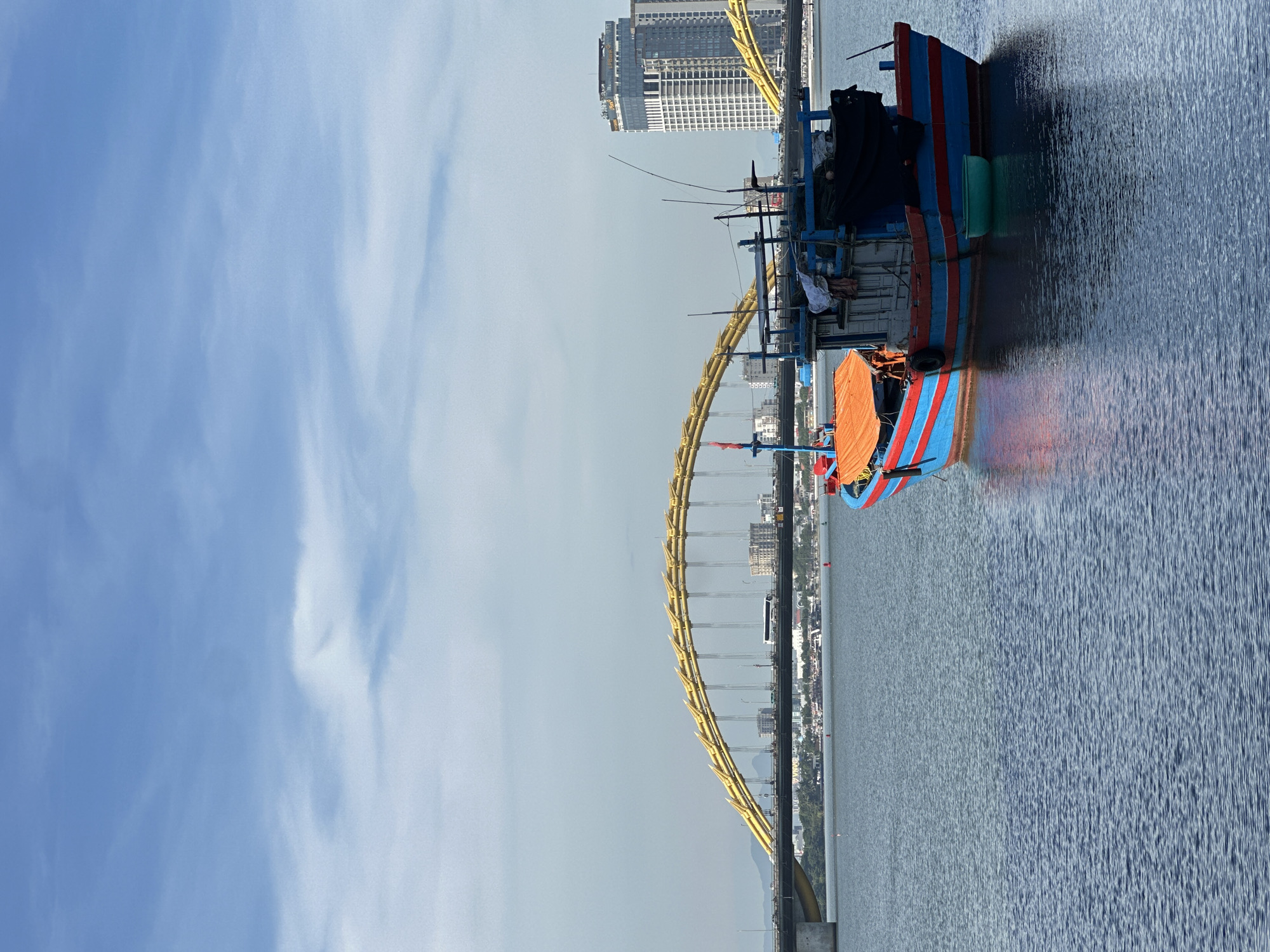 The image captures a bright, sunny day in Da Nang, Vietnam, with a vibrant blue sky and wispy clouds. In the foreground, a colorful fishing boat, painted in bright blue and red stripes, floats on calm, deep blue water. The boat appears weathered and functional, with various equipment and coverings visible on its deck, including an orange tarp and dark fabric. A small, light green basin is affixed to the side of the boat.
In the midground, the iconic Dragon Bridge stretches across the water, its golden, dragon-scale-like structure a prominent landmark. Several cars and a bus are visible traversing the bridge, indicating moderate traffic. Along the riverbank, a cityscape unfolds with various buildings, including a tall, modern high-rise hotel with a distinctive facade. The far background shows a hazy outline of mountains. The overall scene is one of urban coastal life, with a blend of traditional and modern elements.