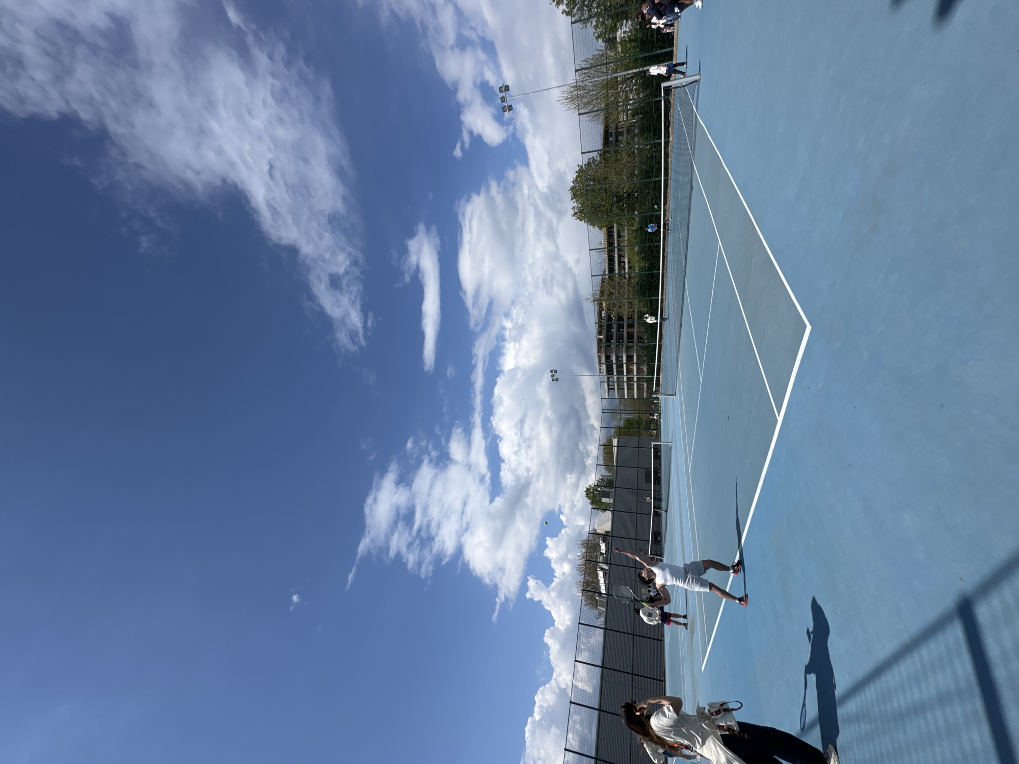 A tennis court is visible under a partly cloudy sky. The court is blue with white lines marking its boundaries. Two players are in action on the court, with one in the midst of serving the tennis ball. A fence runs along the sides of the court, with some spectators standing near the fence. The location is likely in Blagoevgrad, Bulgaria, given the user's prompt.