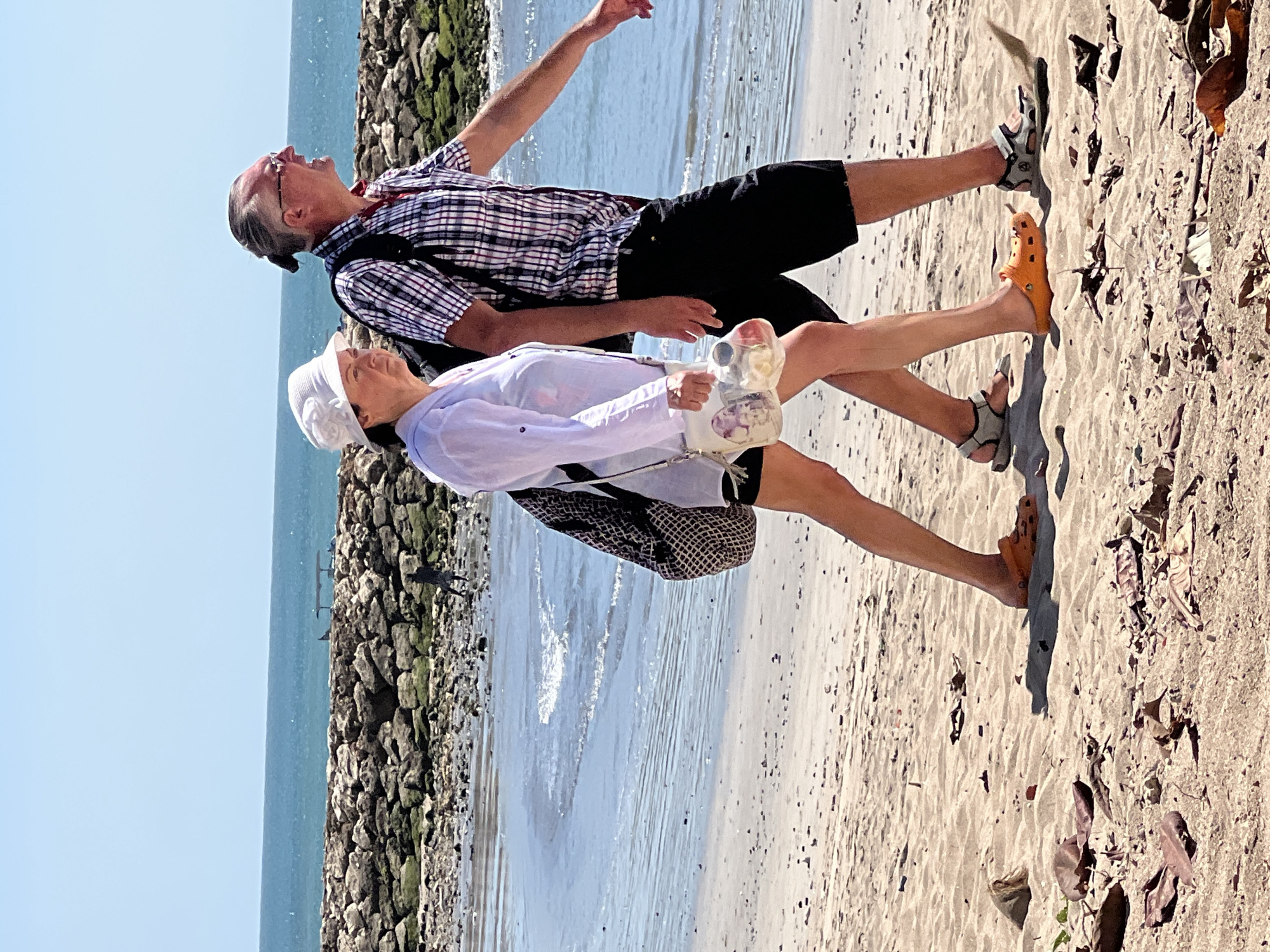 An adult Caucasian couple is walking on a sandy beach in Kuta, Indonesia. The man, on the right, wears a red and white checkered short-sleeved shirt, black shorts, and black sandals. He has graying hair pulled back in a ponytail and is wearing glasses. The woman, on the left, wears a white hat, a white long-sleeved shirt, and dark shorts. She carries a clear bag with floral decorations and a black backpack. They are walking on the wet sand near the ocean's edge. The background shows the ocean, a stone breakwater, and a clear blue sky. The beach is scattered with dry leaves and debris.