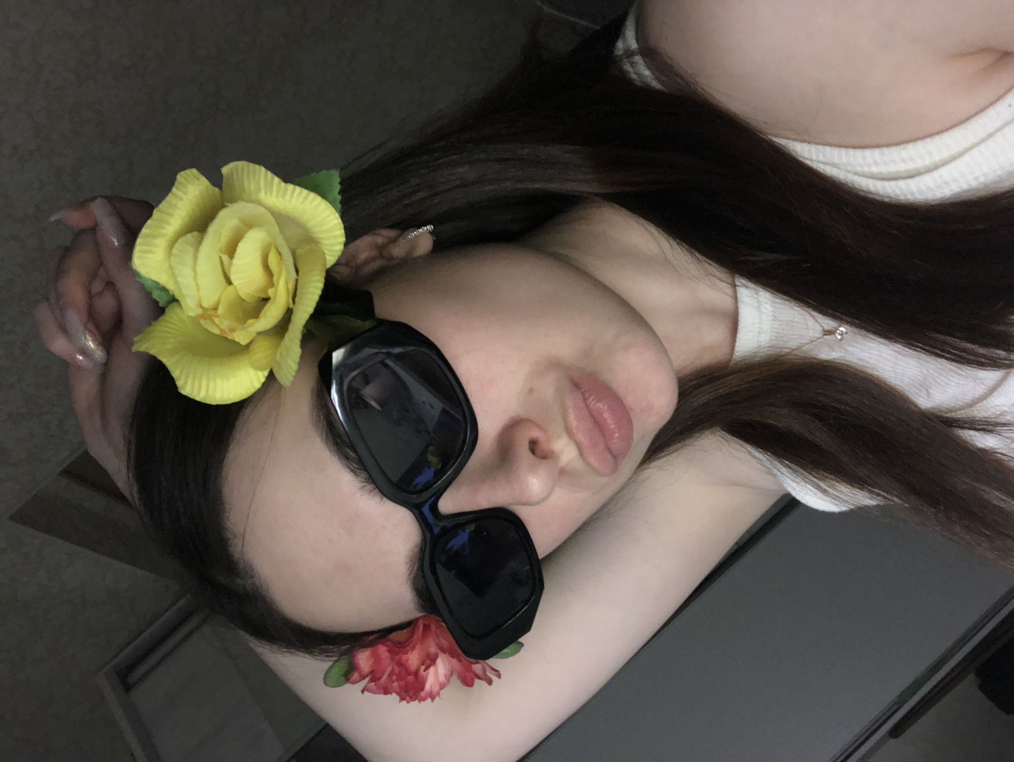 A woman reclines, facing the camera, with a yellow flower behind her ear and a red flower near her sunglasses. Her dark hair is spread out. She is wearing a white top and a necklace. The background is blurred, but appears to be a gray surface. The location cannot be determined from this image.