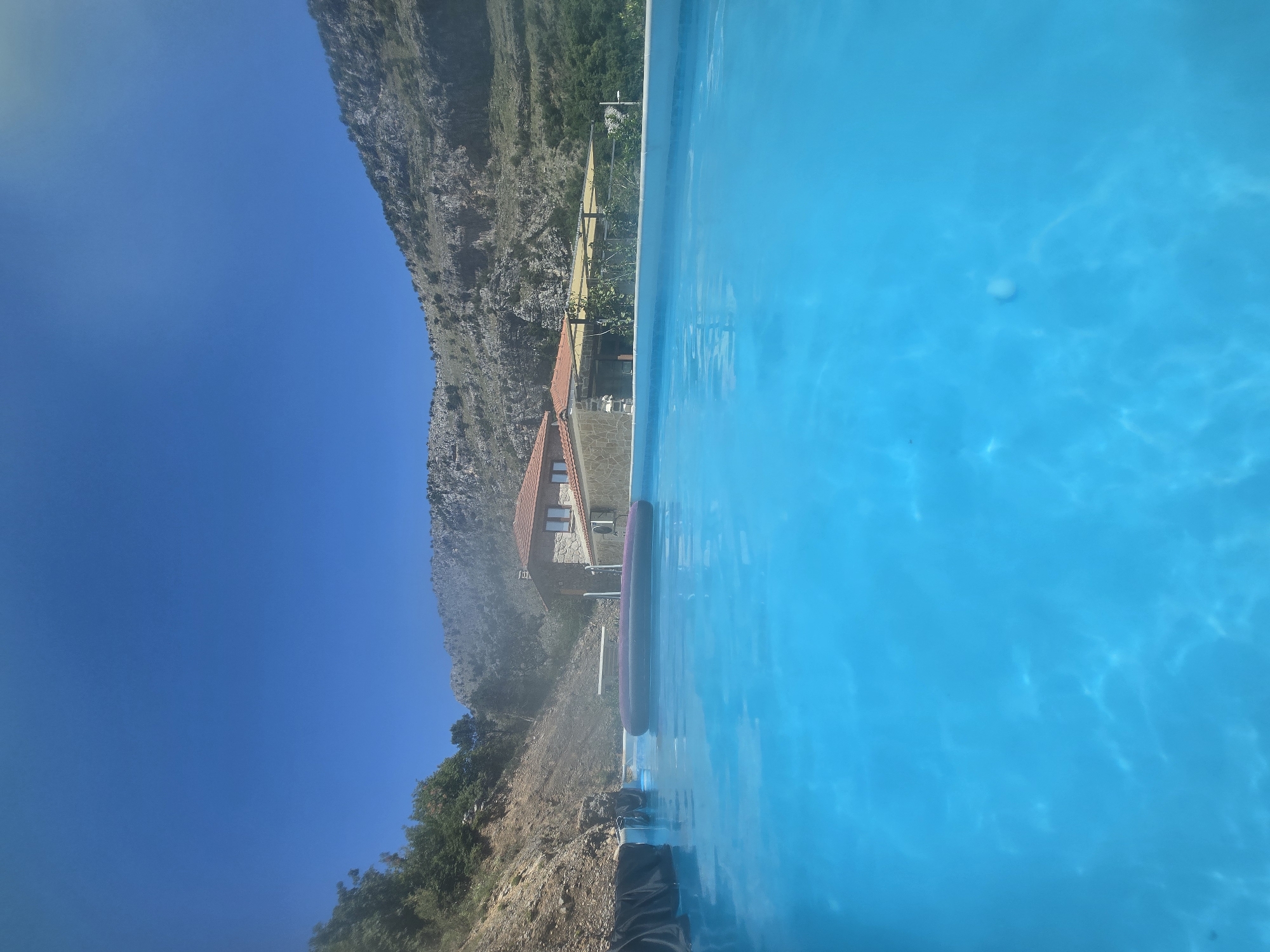 This is a low-angle shot of a bright blue swimming pool with a purple inflatable float in the center. In the background, a stone building with a red tile roof is visible, nestled against a rocky hillside under a clear blue sky. Trees and vegetation can be seen on the left side of the frame, and a portion of a covered patio area is visible to the right of the building. The scene appears to be a serene outdoor setting.