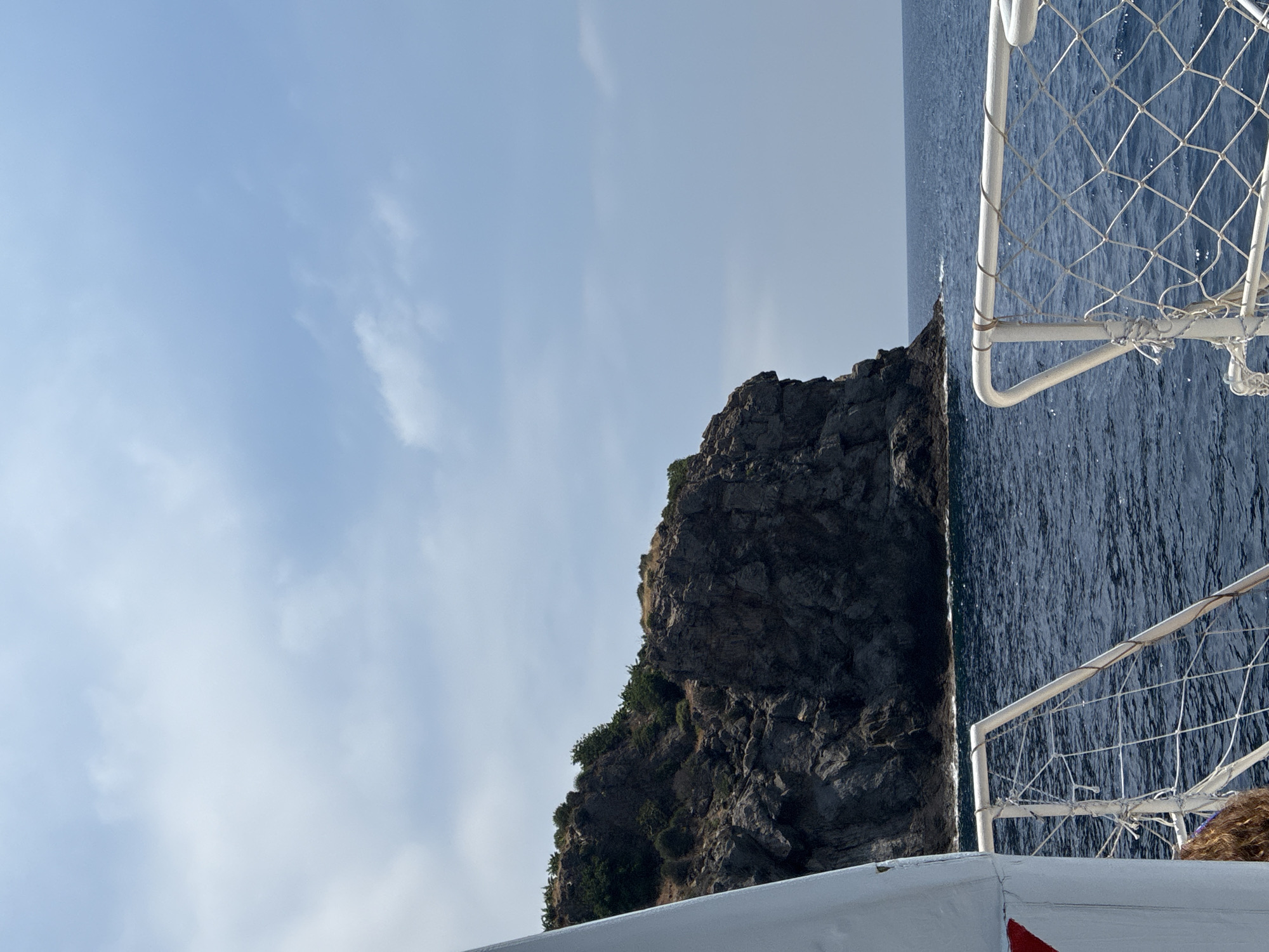 The image depicts a view from a boat sailing on the deep blue Mediterranean Sea, approaching a rugged, rocky island covered in sparse, dry vegetation. The island rises steeply from the water, with a sheer cliff face dominating the right side of the frame. The sky above is a light blue, with scattered white clouds. In the foreground, the white railing of the boat, interwoven with netting, is visible, creating a sense of movement and perspective. A portion of the boat's white hull is also visible on the left side of the image. The scene is likely located off the coast of Gazipaşa, Turkey, given the characteristic landscape of the region.
