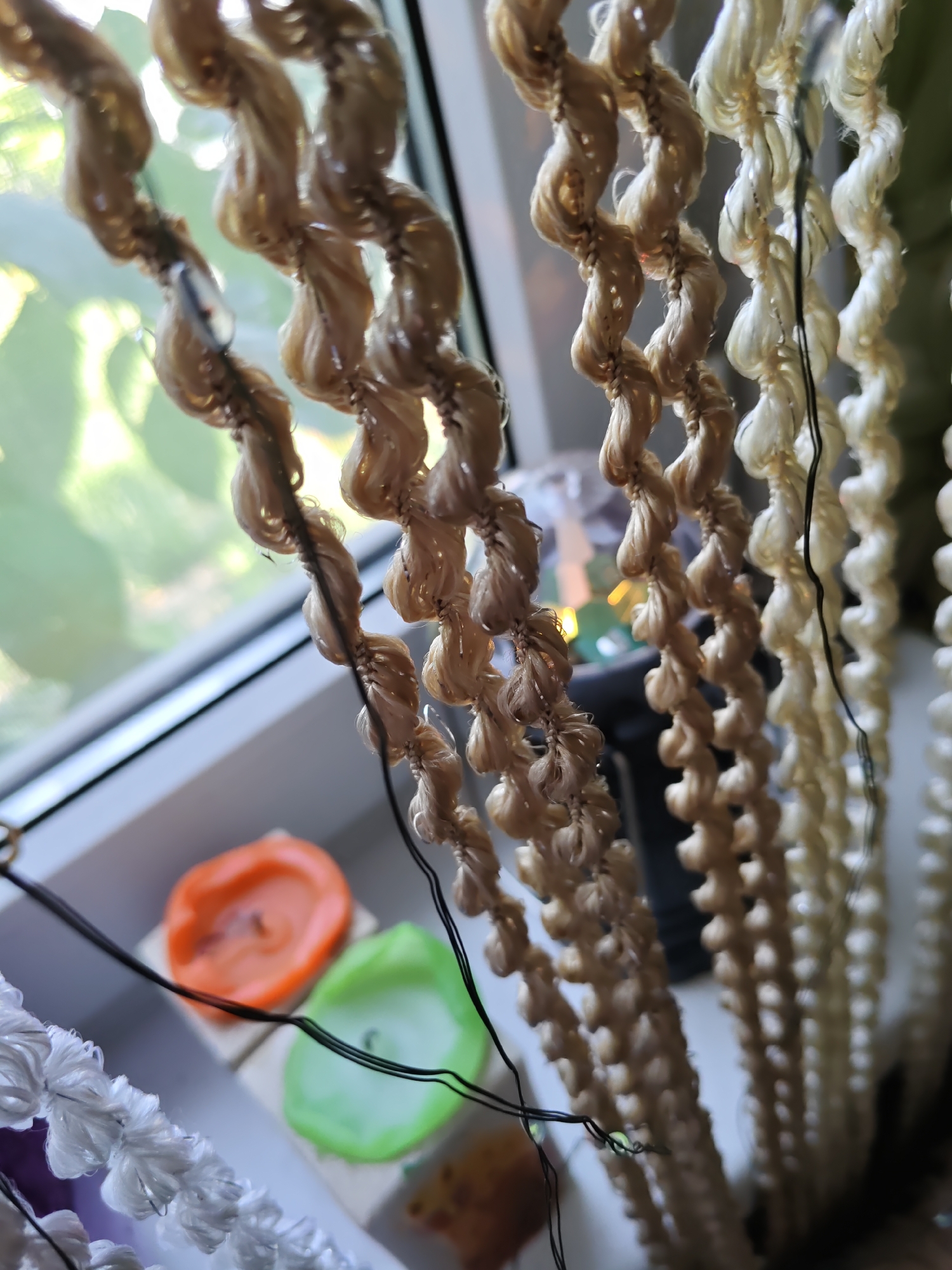 The image shows a close-up view of several strands of twisted rope or cord, hanging vertically. The rope is a light tan or off-white color and has a textured, woven appearance. In the background, out of focus, there is a window with sunlight filtering through, suggesting an indoor setting near a window. Below the ropes and to the left, two small, round candles are visible, one orange and one green. They appear to be set on a white surface. The overall impression is of a decorative arrangement, possibly a craft project or part of a home decor. The image does not provide any information about the city of Rostov-on-Don, Russia, or any specific location.