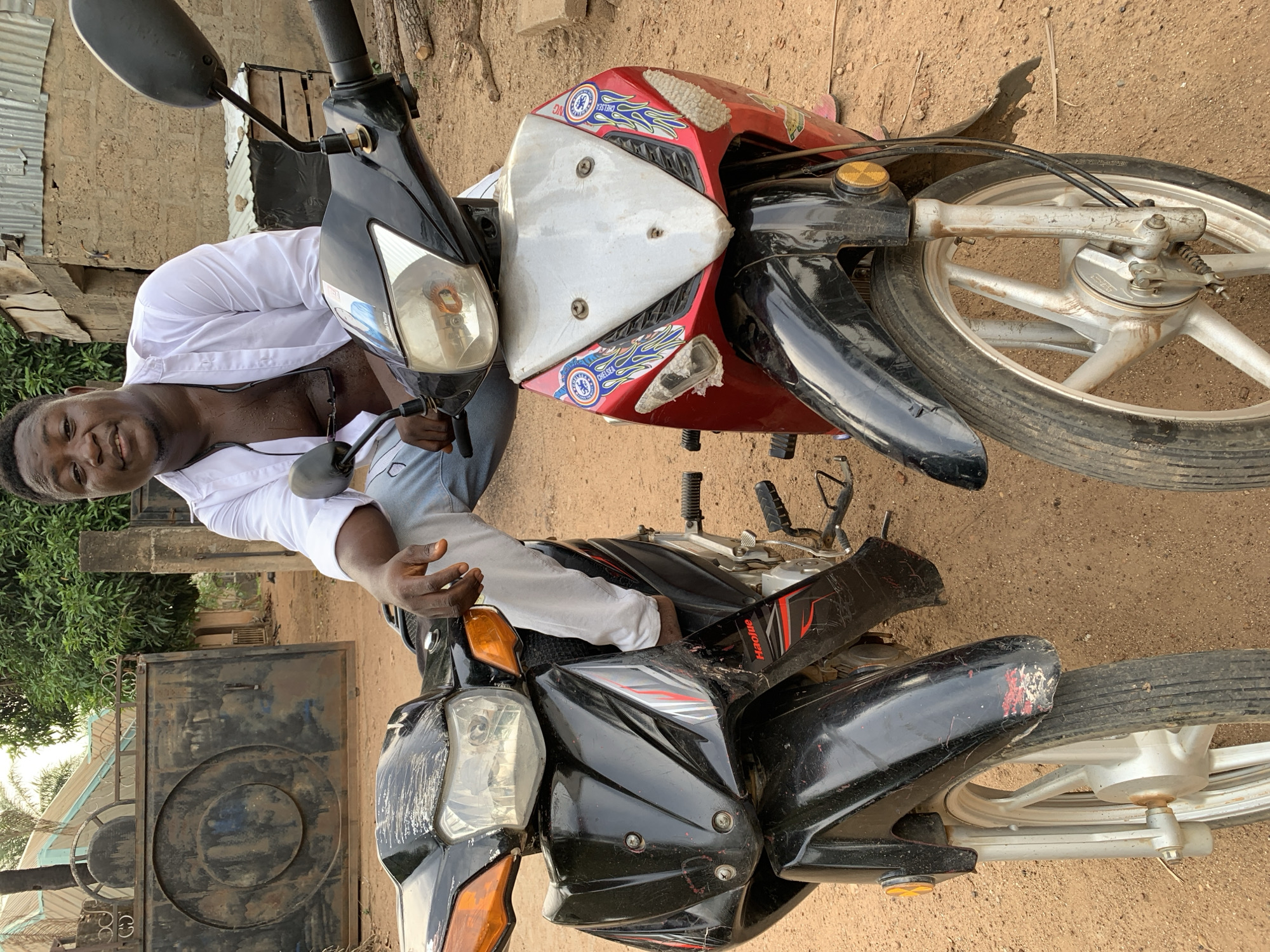 A man smiles directly at the camera, posing outdoors with two motorcycles on a dirt ground in Akwanga, Nigeria. He is dark-skinned, wearing a white long-sleeved shirt unbuttoned to his mid-chest, revealing a sweaty appearance, and light-colored trousers. Sunglasses are hanging around his neck.
He is positioned between two underbone-style motorcycles. The motorcycle on the left (from the viewer's perspective) is predominantly black with red and gray accents, showing numerous scratches and wear. The brand name "Haolue" is visible on its side fairing. The motorcycle on the right is red and white, exhibiting noticeable wear, including peeling stickers and scuff marks. This motorcycle prominently displays several blue "Chelsea" Football Club logos on its fairing. Both vehicles feature silver-colored front wheels.
The background consists of a dirt ground, with various structures and foliage visible. To the left, there is a dark metal gate with a distinctive circular design, along with green trees and parts of a building. To the right, corrugated metal sheets and concrete blocks are discernible. The scene appears to take place during the day under bright light, indicative of warm weather.