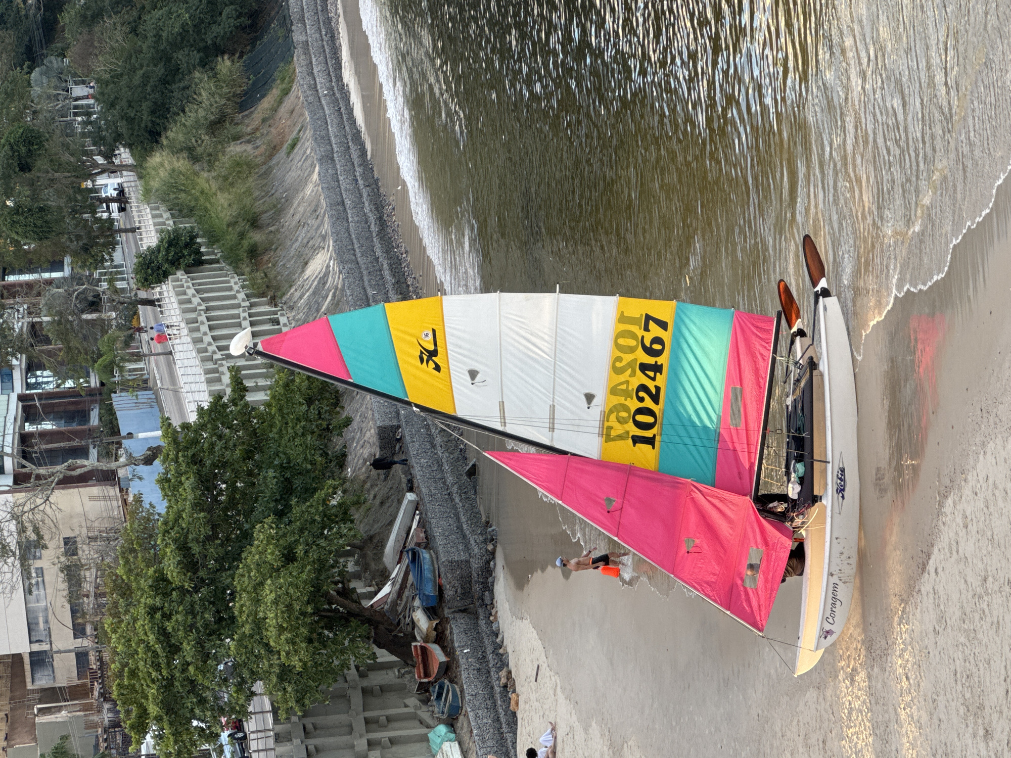 This is a medium-angle, eye-level shot of a Hobie Wave catamaran sailboat on a beach in Niterói, Brazil. The boat has a white hull with the word "Coragem" and a small illustration of a bird in pink lettering on its side. The sail is brightly colored, with sections of pink, teal, yellow, and white. The yellow section displays the number "102467" and the Hobie logo. The sailboat is partially in the shallow water, with its bow on the sand. Two paddles are resting on the side of the boat.
In the background, a sandy beach stretches along the water's edge. Several people are visible on the beach; one person in a blue cap and dark shorts stands in the water near the boat, holding an orange object. Further down the beach, two people are sitting. The beach is backed by a rocky embankment and a retaining wall. A few small boats are piled on the embankment.
Beyond the embankment, a street runs parallel to the beach. Cars are visible on the street, and pedestrians are walking along a paved walkway next to the street. Buildings, including a modern structure with large windows, are situated behind the walkway and trees. The scene appears to be in a coastal urban area, likely a public beach with recreational activities. The lighting suggests it is late afternoon or early morning, with the sun casting long shadows.
