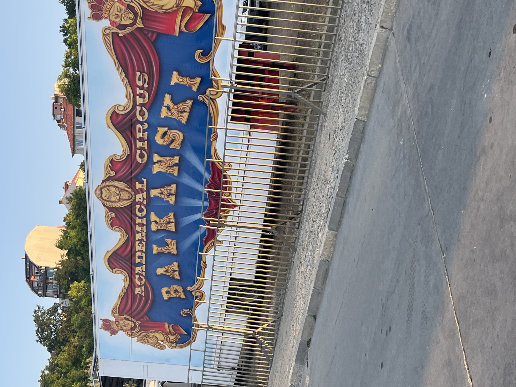 The image captures a close-up of a circus trailer with "ACADEMICAL CIRCUS BALKANSKI" painted in large, ornate letters across its side. The artwork features a regal design with crowns, lions, and red drapery. The trailer is parked outdoors, with a metal fence in the foreground and a cobblestone area behind it. In the background, buildings and trees suggest an urban or semi-urban setting. The sky is clear and blue. The location is identified as Nesebar, Bulgaria.