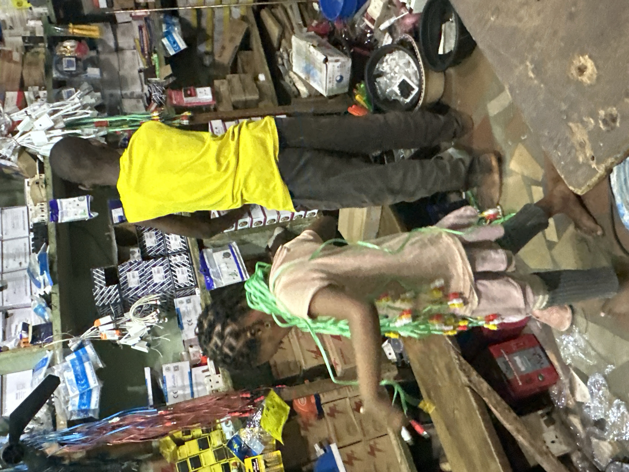In a cluttered indoor market stall in Narayi, Nigeria, a young girl with braided hair, adorned with a long green string of colorful beads, stands facing left, her body angled slightly towards the viewer. She wears a light pink t-shirt and dark grey leggings. Her right arm is extended, appearing to interact with items on a wooden shelf in front of her, while her left arm is bent at the elbow. Another individual, a dark-skinned male wearing a bright yellow sleeveless shirt and dark grey pants, stands to her right, facing away from the camera and towards the shelves behind the girl.
The stall is densely packed with various goods. Shelves are laden with boxes, electrical components, and assorted items. In the foreground, a wooden surface, possibly a counter or table, is visible. The floor appears to be tiled with geometric patterns. To the right of the male figure, two plastic bowls are present, one containing white material and the other empty. Various cables and smaller packaged items are displayed on hooks and shelves throughout the stall. The overall impression is one of a busy, disorganized retail environment.