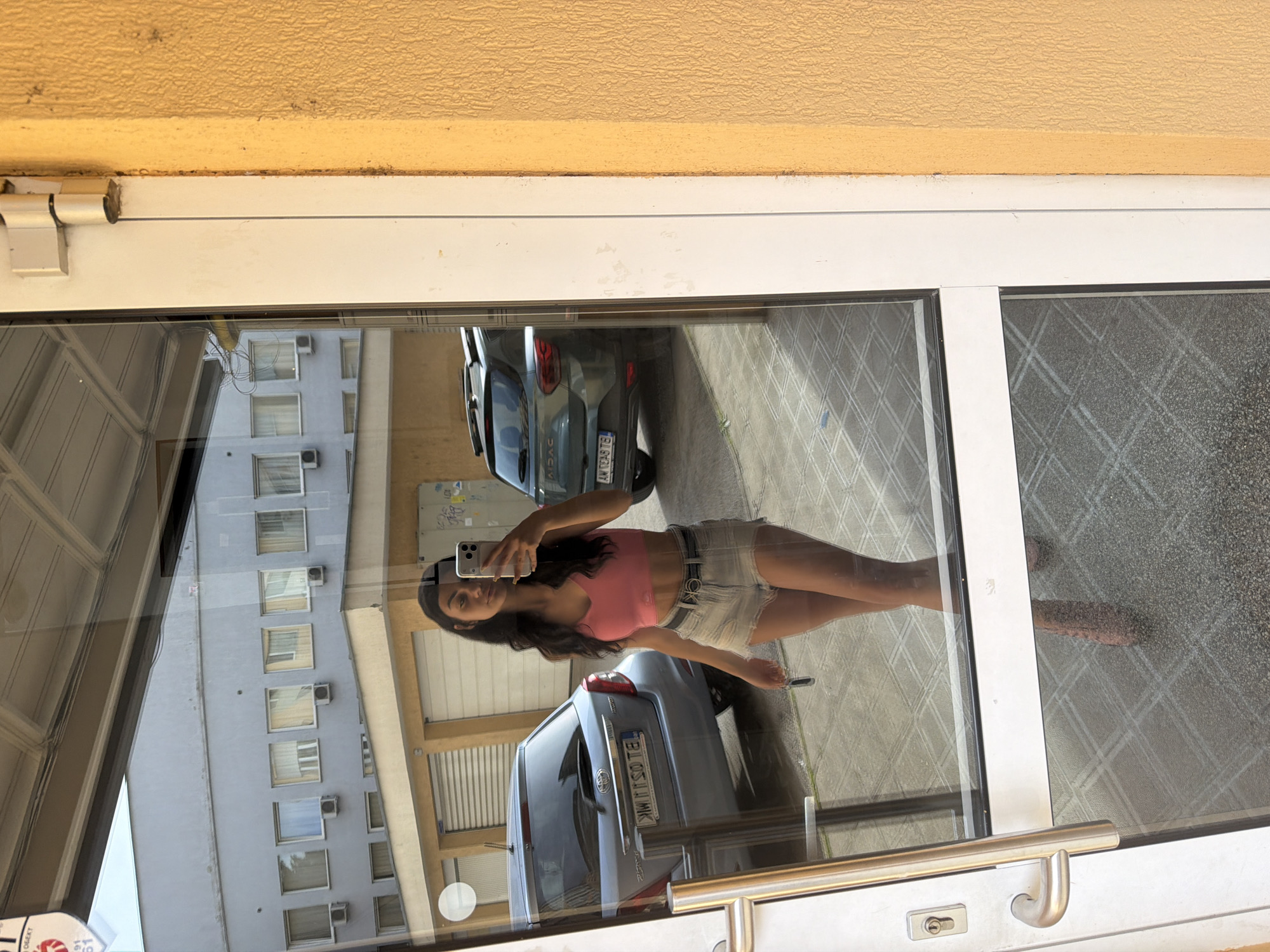 A young woman with dark, wavy hair takes a mirror selfie in front of a glass door. She is wearing a pink crop top and denim shorts, with high-heeled shoes. She is holding a phone in her right hand and a set of keys in her left. Behind her, reflected in the glass, are two cars parked in what appears to be a parking lot. The building in the background is light gray with rows of windows. The scene suggests a casual, urban setting. The woman appears to be in Veliko Tarnovo, Bulgaria, as indicated by the license plates on the cars.