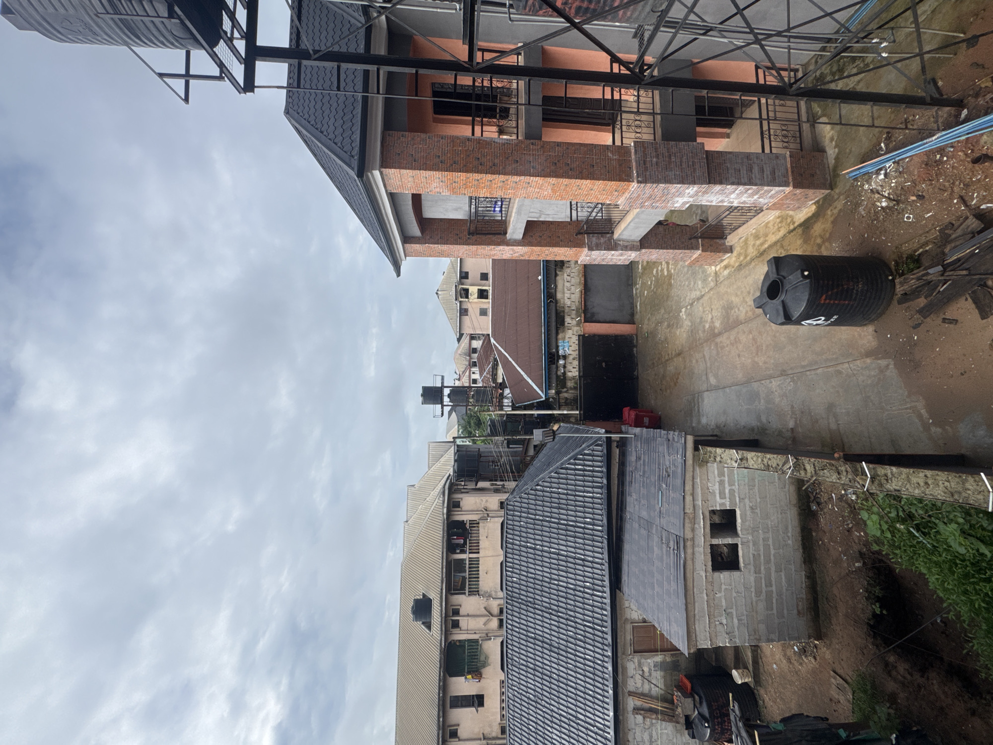 The image depicts an overcast sky above a built-up area in Ihiagwa, Nigeria. In the foreground, to the right, is a multi-story building under construction with an orange facade and brick pillars, featuring balconies with black metal railings. Adjacent to this building, to the left, is a smaller, partially constructed building made of grey concrete blocks with two square openings. Further back, a long, pale pink building with multiple windows and balconies is visible, with large black water tanks on its roof and on a raised platform behind it. The ground is a mix of paved areas and dirt, with scattered debris. Another black water tank is positioned on the ground between the buildings. The overall scene suggests a developing urban or semi-urban environment.