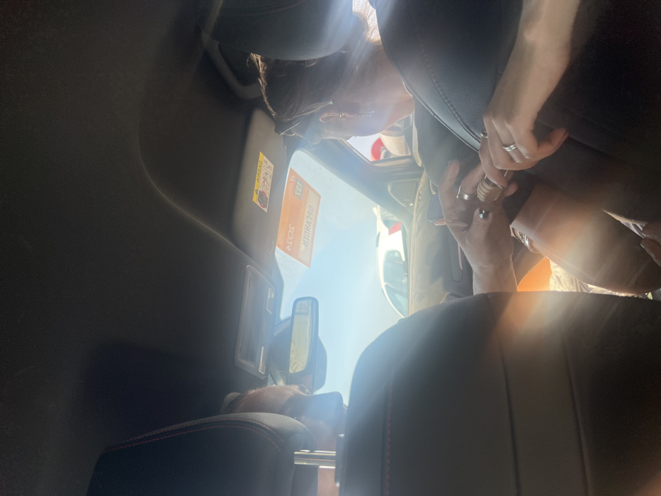 The image captures the interior of a vehicle, looking forward from a rear seat, under bright daylight in Parikia, Greece. Two individuals occupy the front seats. The driver, a male, is seated on the left, visible from behind a dark headrest with red stitching. His left hand, adorned with a ring, is extended towards the center. The front passenger, a female, is seated on the right, also visible from behind a similar headrest. She wears sunglasses on her head and earrings, and multiple rings are visible on her left hand. Her right hand holds a small, cylindrical object of light-colored, possibly wooden material, featuring metallic accents. The male's left hand is engaged with this object, their fingers interlocked as if in the act of opening or closing it. Through the front windshield, a clear blue sky is visible above the rear of a white vehicle and a dark-colored vehicle, indicating an outdoor setting, likely a road or parking area. The vehicle's sun visor on the passenger side displays an "AIRBAG" warning label and a rectangular orange sticker with Greek text, identifying it as from the "Hellenic Republic / Ministry of Citizen Protection / Police" with "SOS" and the emergency number "100." Sunlight causes noticeable lens flares within the cabin.