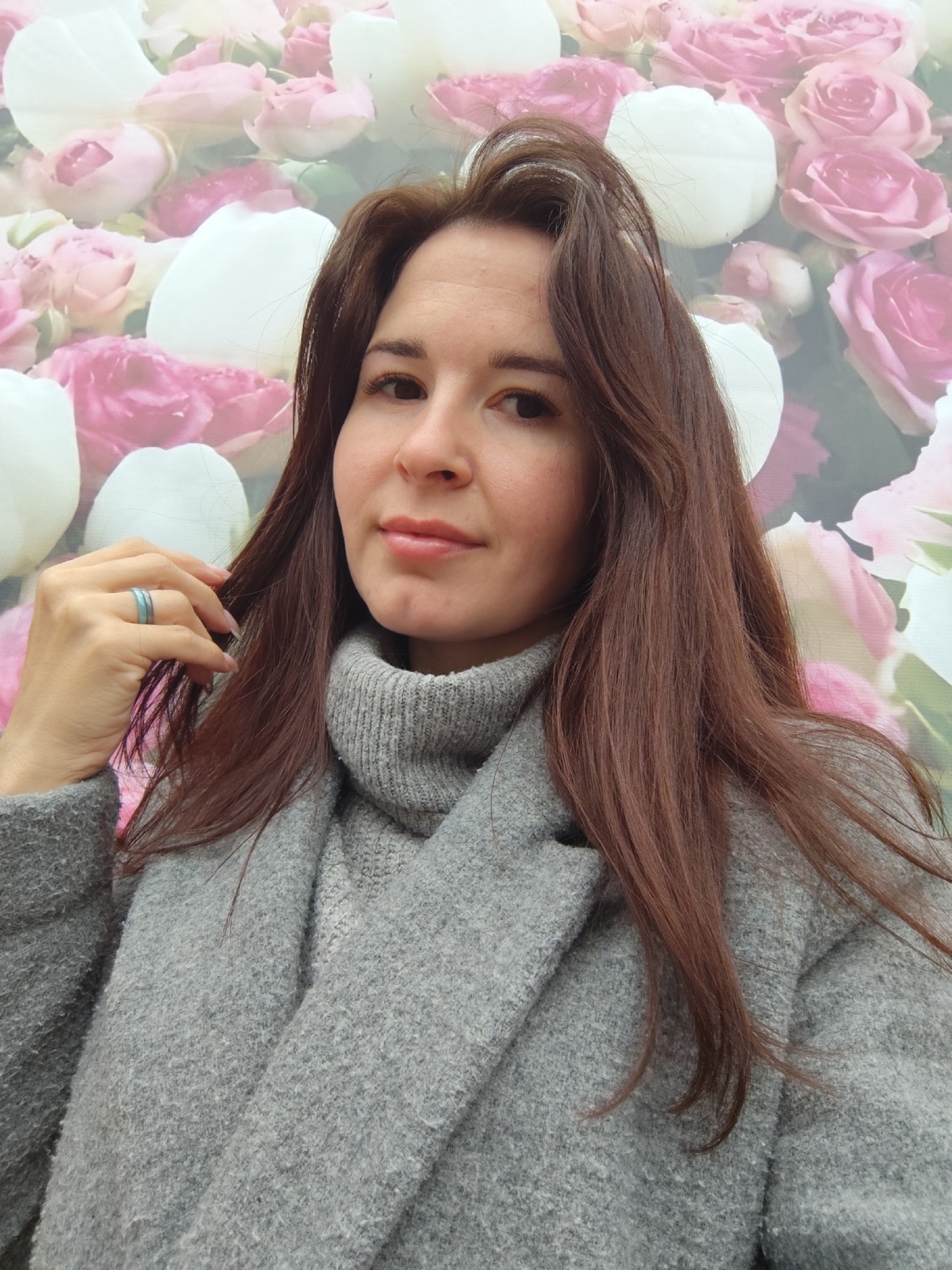 A woman is centered in the frame, wearing a grey turtleneck sweater and a grey coat. Her auburn hair falls to her shoulders. She wears a ring on her left ring finger. The background is a floral design with pink and white roses. The photo may have been taken outdoors, in Фоки, Russia.
