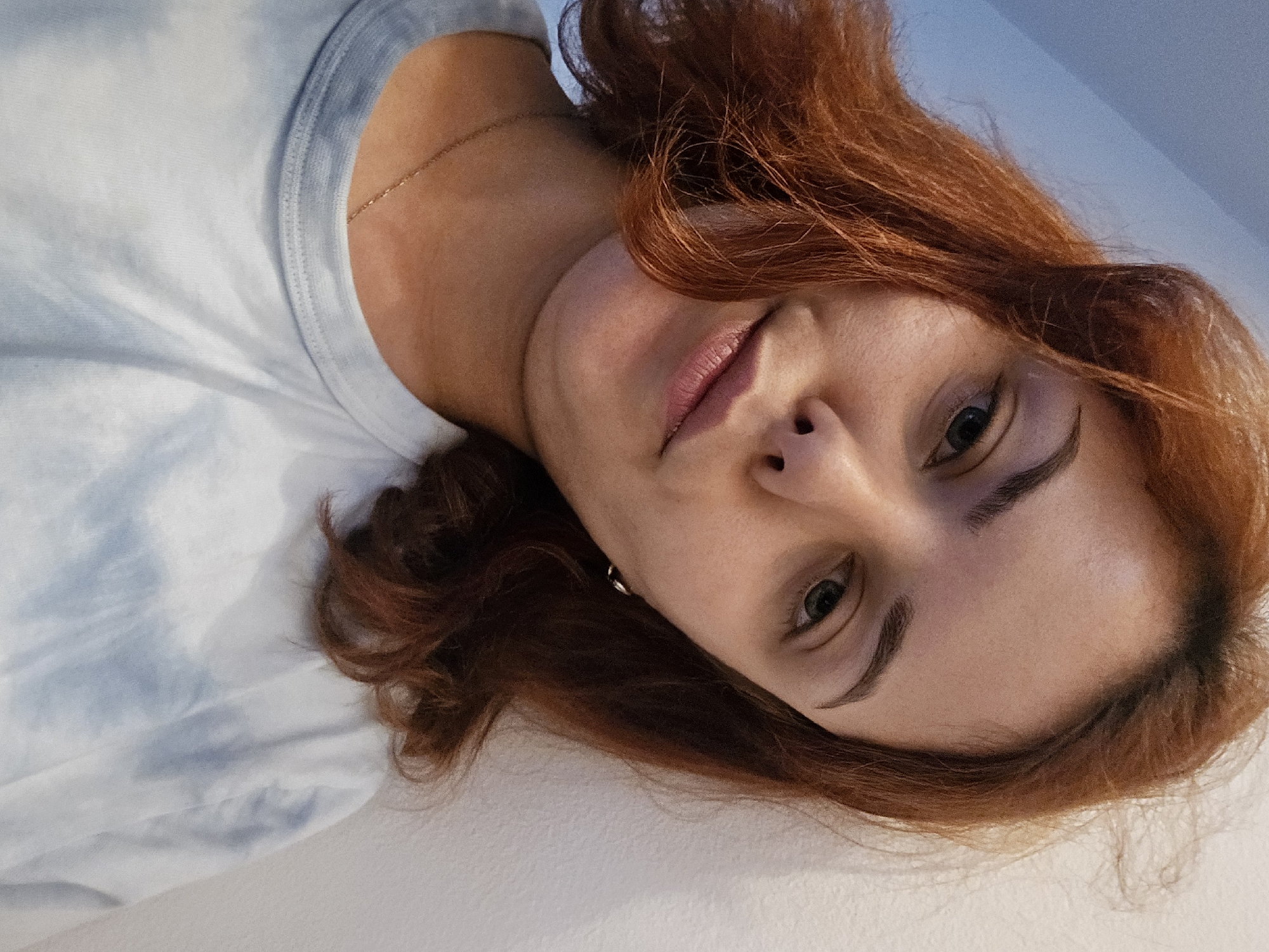 A young woman with reddish-brown hair and green eyes is captured in a close-up, angled selfie. She is leaning against a white wall, with the wall and a pale blue ceiling visible in the background. She is wearing a light blue tie-dye t-shirt and a delicate gold chain necklace. Her expression is neutral, with a slight hint of a smile. The lighting is soft, creating subtle shadows on her face. The overall atmosphere is casual and intimate.