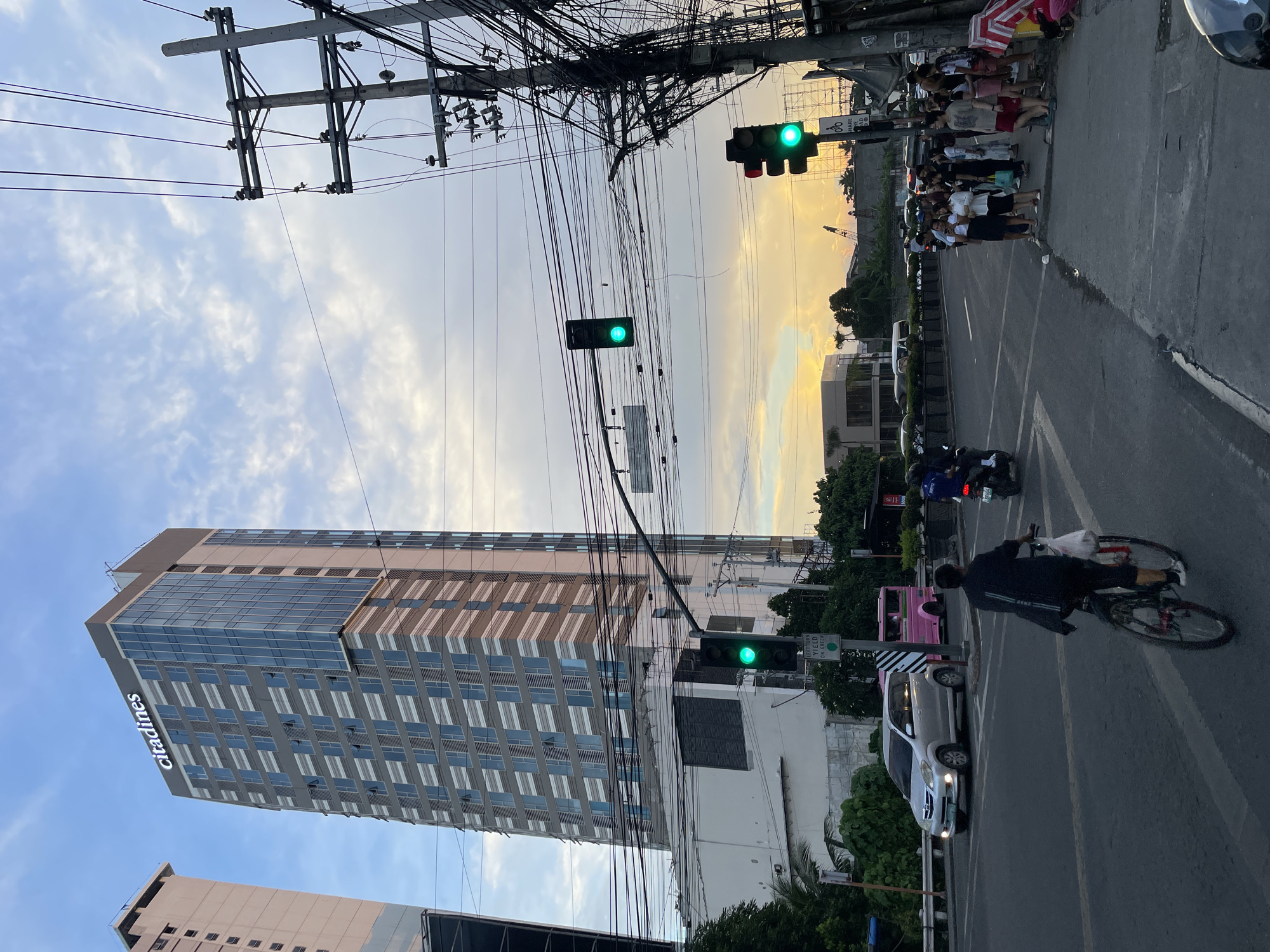 The image captures a street scene in Davao City, Philippines, likely during a sunset given the warm hues in the sky. A tall, modern building with the "Citadines" logo is prominently visible on the left, dominating the skyline. The building has a patterned facade with many windows. In the foreground, a busy intersection is depicted with traffic lights, vehicles, and pedestrians. A silver car and a pink jeepney are among the vehicles. A cyclist is riding on the right side of the road, facing away from the camera. A group of people are gathered on the sidewalk near a utility pole laden with numerous electrical wires. The overall atmosphere suggests an urban environment with active street life.
