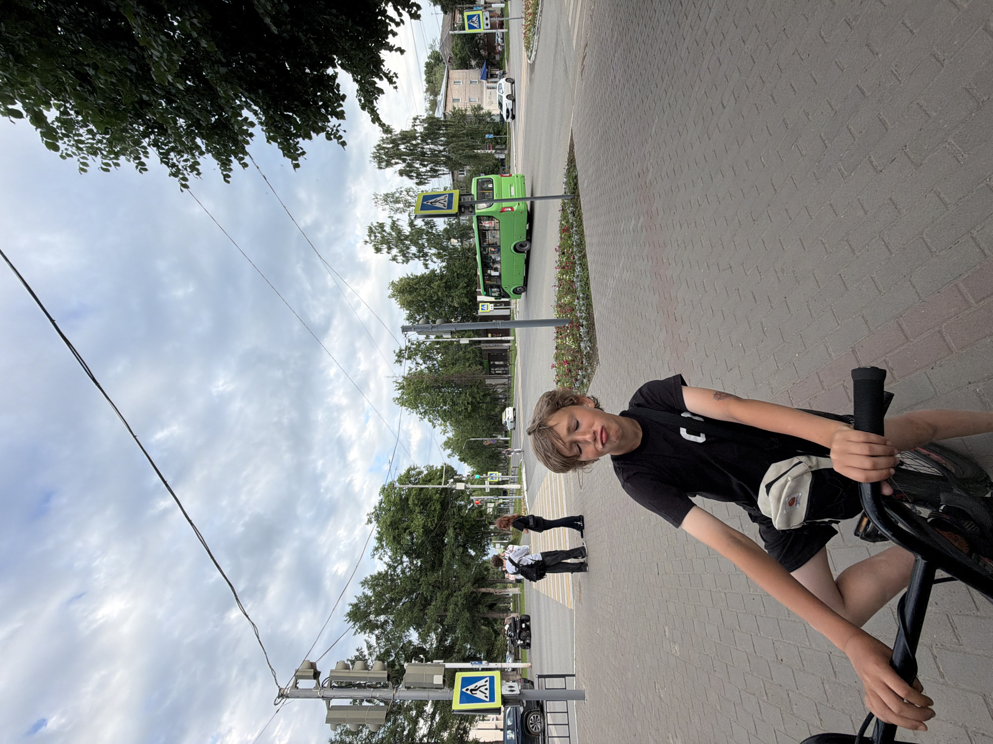 This media file depicts a scene from Chaikovsky, Russia, with a young person on a bicycle in the foreground. The person is wearing a black t-shirt and shorts, with a light-colored fanny pack around their waist. Their eyes are closed and their head is tilted back, suggesting a moment of relaxation or playfulness. The bicycle is black and appears to be a BMX style.
In the background, a green bus is visible on a paved road, approaching a pedestrian crossing. The crossing is marked with white stripes and a blue pedestrian crossing sign is prominently displayed. There are also other people waiting at the crossing. The setting appears to be a street in a town, with trees and buildings lining the road. The sky is partly cloudy, indicating daytime. The overall mood seems casual and summery.