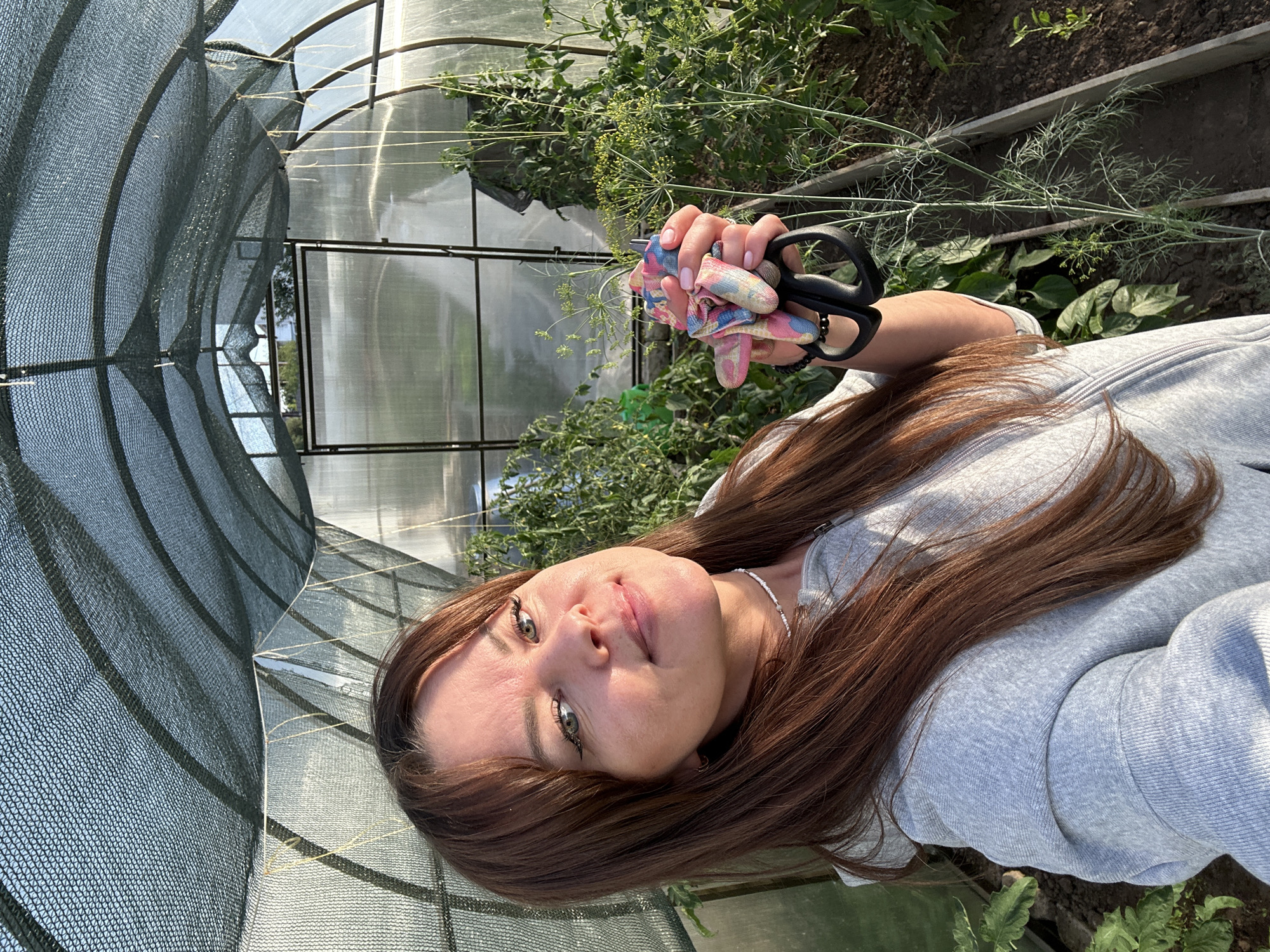 In a greenhouse located in Chaikovsky, Russia, a woman with long, reddish-brown hair and fair skin is posing for a selfie. She is wearing a grey hooded jacket and a thin beaded necklace. Her left hand holds a pair of black-handled scissors and a colorful cloth, possibly gardening gloves, in front of a blurry background of green plants, some of which appear to be tomatoes and dill. The greenhouse structure is visible, with a netted canopy overhead and clear plastic sheeting on the sides. The lighting suggests it is either early morning or late afternoon, casting long shadows and warm tones on the scene.