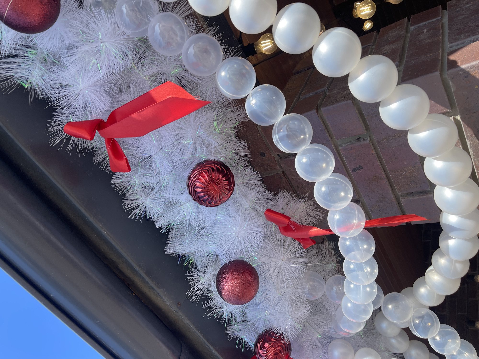 A low-angle shot depicts a holiday decoration. A white, spiky garland is adorned with red spherical ornaments and red ribbons. A string of clear and white plastic orbs is draped nearby. The setting appears to be a building exterior, with brickwork and the edge of a dark-colored overhang visible. The sky is blue. The location is unknown.