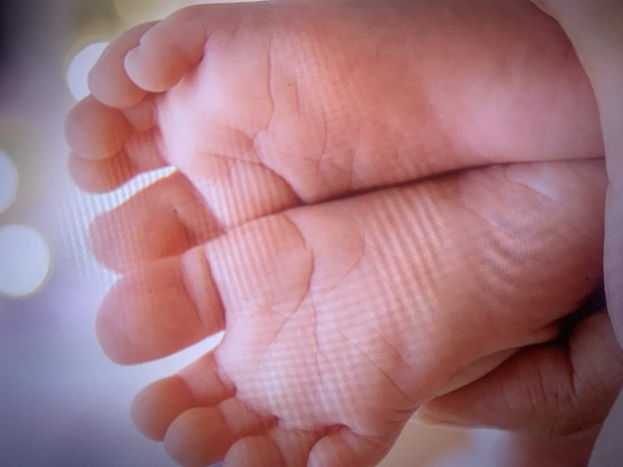 The image is a close-up, abstract shot of two tiny baby feet. The feet are fair-skinned and appear to be those of a newborn. The soles of the feet, with their delicate creases and lines, are prominently displayed. The fingers of an adult hand are visible, gently cradling and supporting the baby's feet, suggesting a moment of tender care and affection. The background is soft and blurred, with diffused circular lights, possibly indicating an indoor setting with soft lighting or a shallow depth of field. The overall impression is one of intimacy, fragility, and love. There is no visible text. The scene evokes a sense of peace and new beginnings.