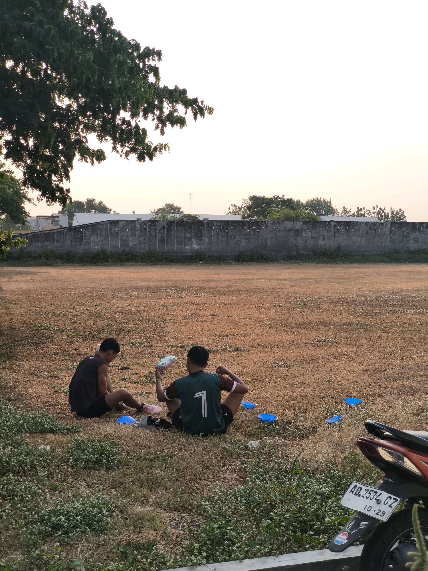 Two young males are sitting on dry grass on a sports field in Desa Sindon, Indonesia. One male, wearing a black tank top and shorts, is looking down at something in his hands. The other male, wearing a green jersey with the number "1" on the back and black shorts, is holding a plastic bag of water bottles. In front of them are several blue cones, likely used for drills. The field is bare and dry, with a concrete wall and buildings in the background. A motorcycle is parked to the right of the frame, with a license plate reading "AD.2534.CZ 10-29". The scene is illuminated by the soft light of what appears to be either sunrise or sunset.