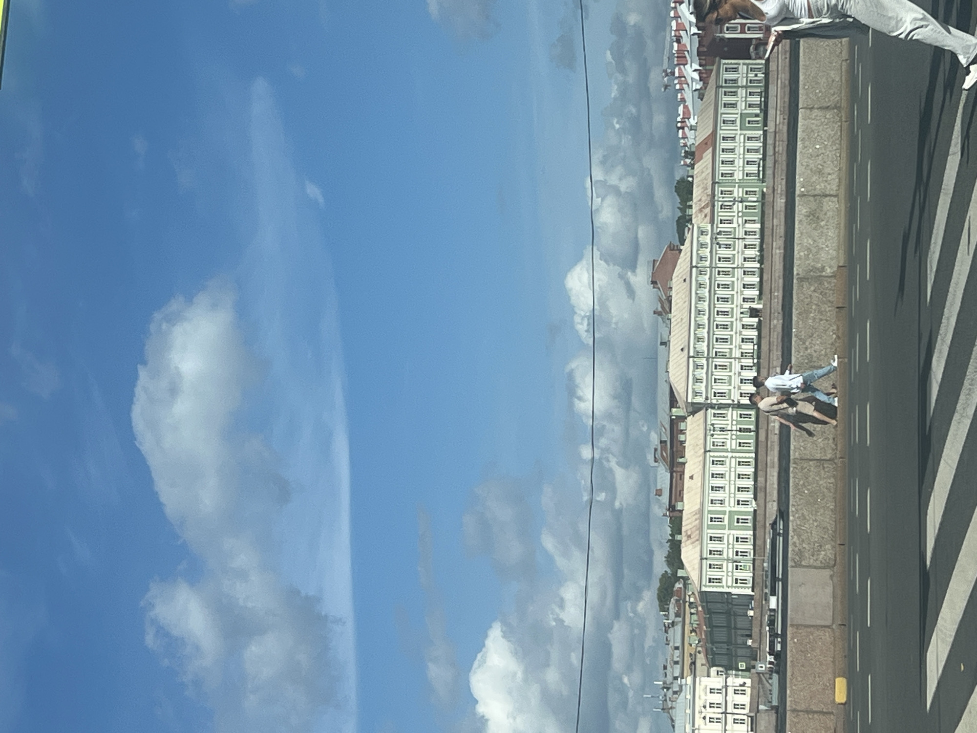 An eye-level shot shows a sunny day in an urban environment. The sky is a vibrant blue with scattered white clouds. In the foreground, a paved road with white dashed lines and a pedestrian crossing is visible. A woman in a white top and gray pants walks away from the camera on the right side of the frame. To her left, two figures, one in a tan shirt and shorts and the other in a white shirt and blue jeans, walk side-by-side across the pedestrian crossing. A concrete barrier separates the road from a lower level. Beyond the barrier, a wide body of water is seen with a white boat or ferry. On the far side of the water, a row of historic buildings with green facades and white window trim line the waterfront. The buildings have red roofs and multiple stories. In the background, more buildings and a few trees are visible. A thin black wire stretches horizontally across the middle of the frame.