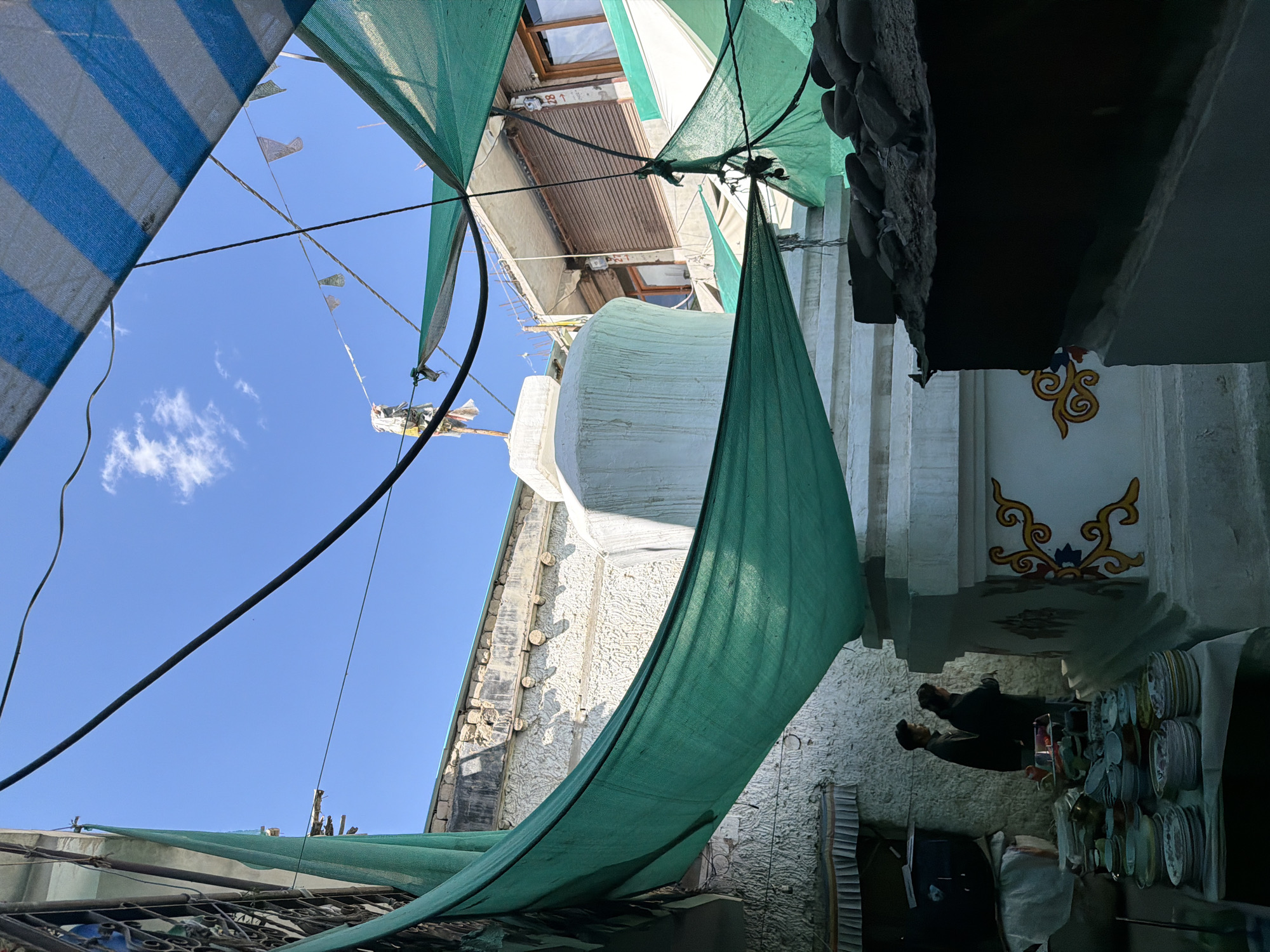 In Leh, India, a white stupa stands prominently amidst a cityscape partially obscured by overhead green and blue striped tarps. Two individuals, one man and one woman, stand near the base of the stupa, facing away from the viewer. In the foreground, a table is laden with stacks of colorful ceramic plates and bowls, suggesting a marketplace or a domestic setting. The stupa itself is intricately decorated with yellow and red swirling patterns. The sky above is a clear blue, with a few wispy clouds. The architecture in the background suggests a densely built urban environment.