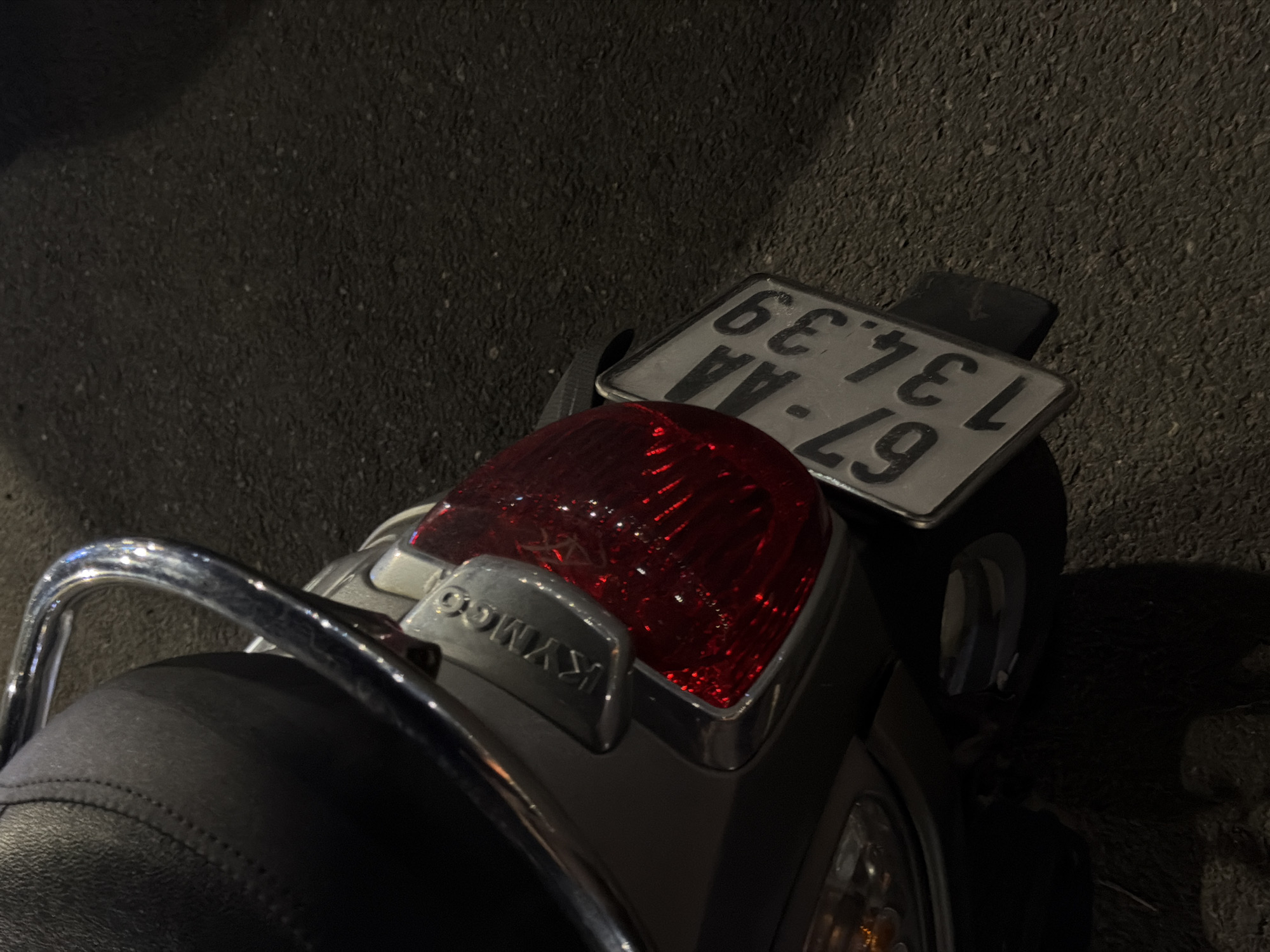 The image shows the rear of a dark-colored scooter parked on asphalt at night. The license plate, with text reading "67-AA.134.39", is partially visible. A red taillight with chrome trim is prominent. The scooter has a black seat and a chrome grab handle. The background is dark asphalt, suggesting an outdoor setting, possibly a street or parking lot. The lighting is dim, indicating it is nighttime. There are no people visible in the image. The scene is static, with the scooter parked. The location context is identified as Đà Nẵng, Vietnam, which is consistent with the scooter's design and license plate format.