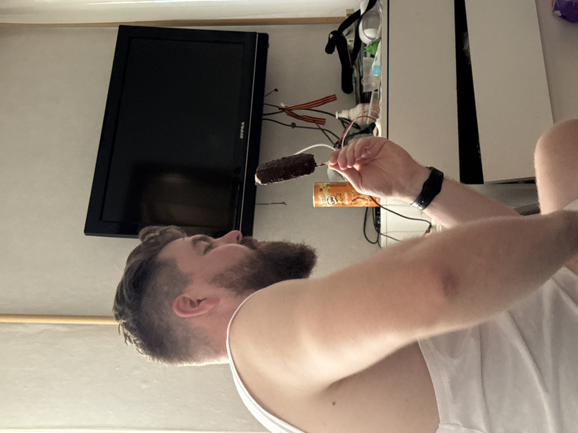A medium shot captures a man in a white tank top from the chest up, seated and facing to the left, holding a chocolate-covered ice cream bar on a stick. He has a full beard and short, dark hair, with the side of his head visible. His right hand, holding the ice cream, is extended towards the viewer. The background includes a black television screen mounted on a wall, with various items on a surface below it, including a Pringles can, wires, and a bottle. The scene appears to be indoors, possibly a bedroom or living area. The man is wearing a black fitness tracker on his left wrist. The overall lighting suggests it is daytime.