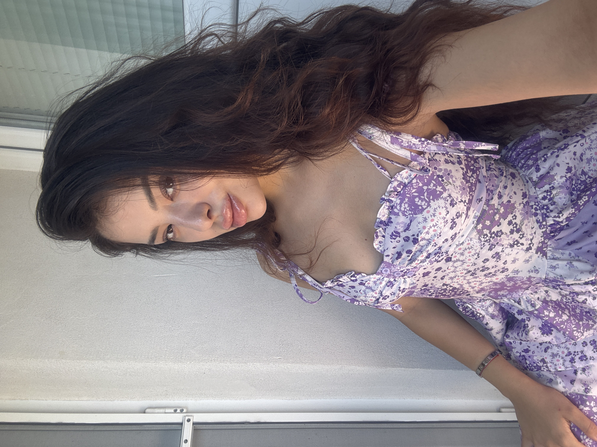 A young woman with long, dark brown hair is the central subject of the image. She is wearing a purple and white floral print dress with thin straps. A bracelet adorns her left wrist.
She appears to be indoors, possibly near a window. The background consists of a white wall and a window frame, suggesting an apartment or room.
There is no verifiable information from the image to suggest a location in Plovdiv, Bulgaria. The scene is generic and could be located anywhere.