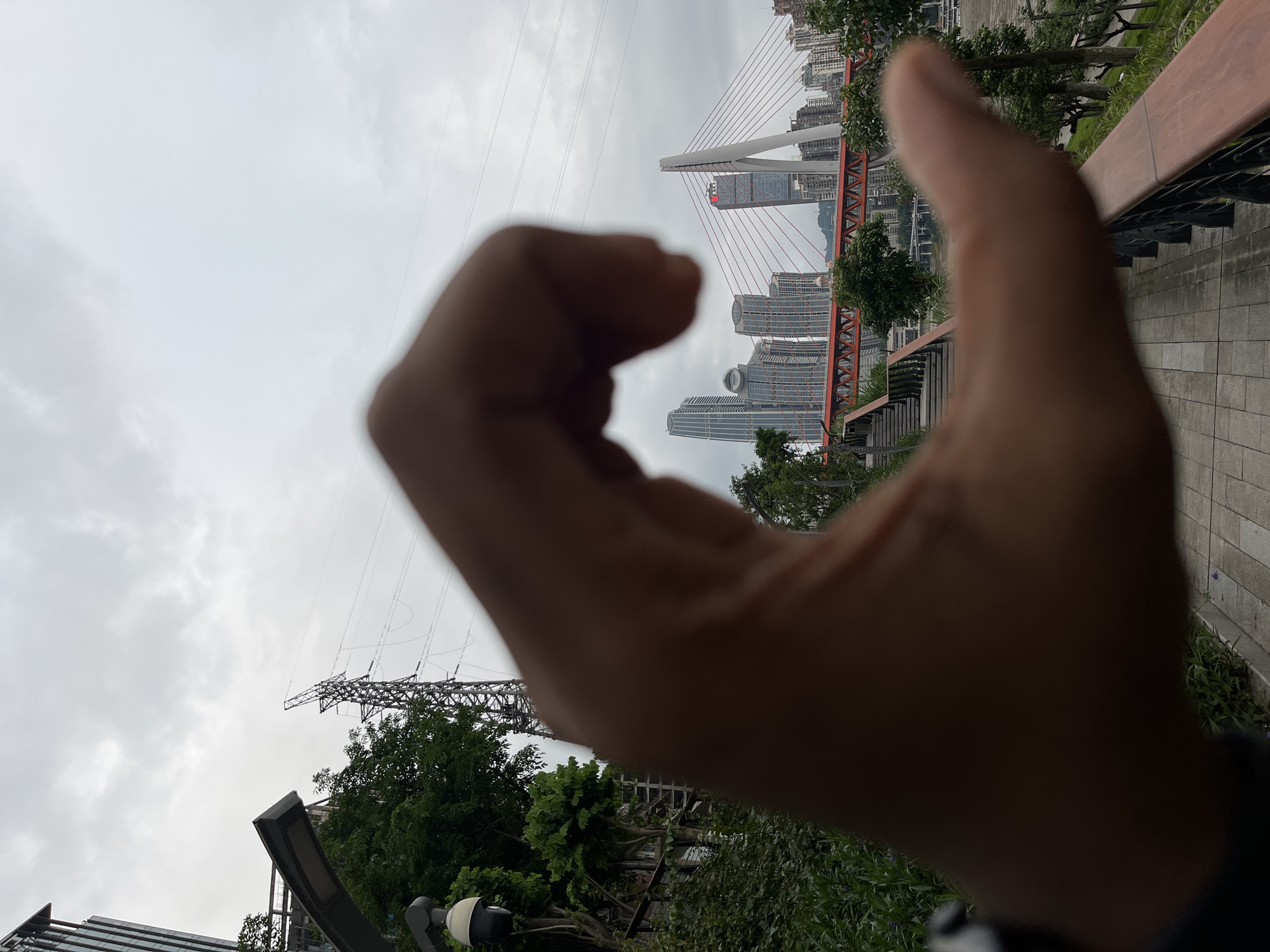 A person's hand is positioned in the foreground, forming an "O" shape with the fingers. The background features a cityscape, including a cable-stayed bridge with red supports, and modern high-rise buildings. Electrical towers are visible on the left side of the image. The scene is 江北城街道, China. The sky is overcast. The photograph is taken outdoors.
