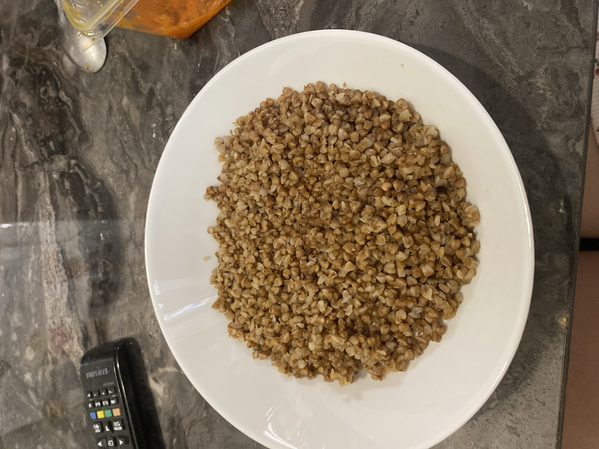 The scene depicts a white plate filled with buckwheat grains. The plate is positioned on a dark, possibly stone-like, surface. A Samsung remote control lies next to the plate on the same surface. A container partially visible above the plate holds an orange-colored substance, potentially sauce or another food item. The context of Chaikovsky, Russia is provided, however, there are no recognizable markers to verify this geographic information.