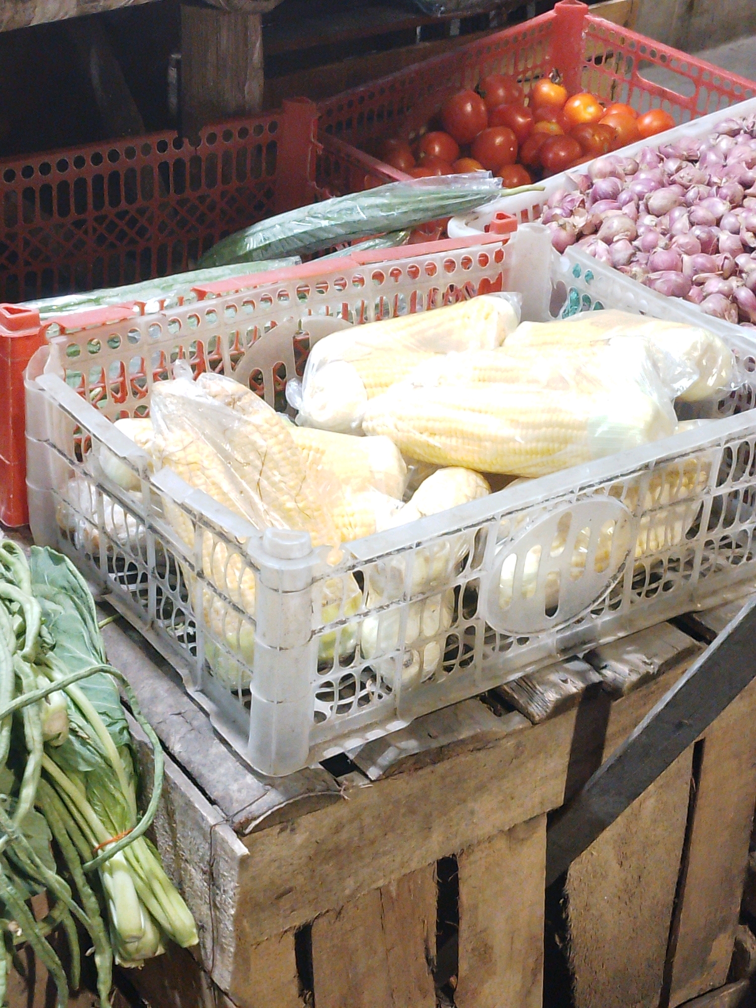 A high-angle medium shot captures a market stall in Kertosono, Indonesia. In the foreground, a bundle of green long beans, tied with a red rubber band, rests on a wooden surface. To the right, a white plastic crate holds several ears of corn, some wrapped in plastic. Behind this, a red plastic crate is filled with tomatoes and a white plastic crate contains a pile of small, purple shallots. In the background, another red plastic crate is partially visible, holding what appear to be cucumbers or similar green vegetables. The scene is illuminated by ambient light, casting shadows across the wooden structures.