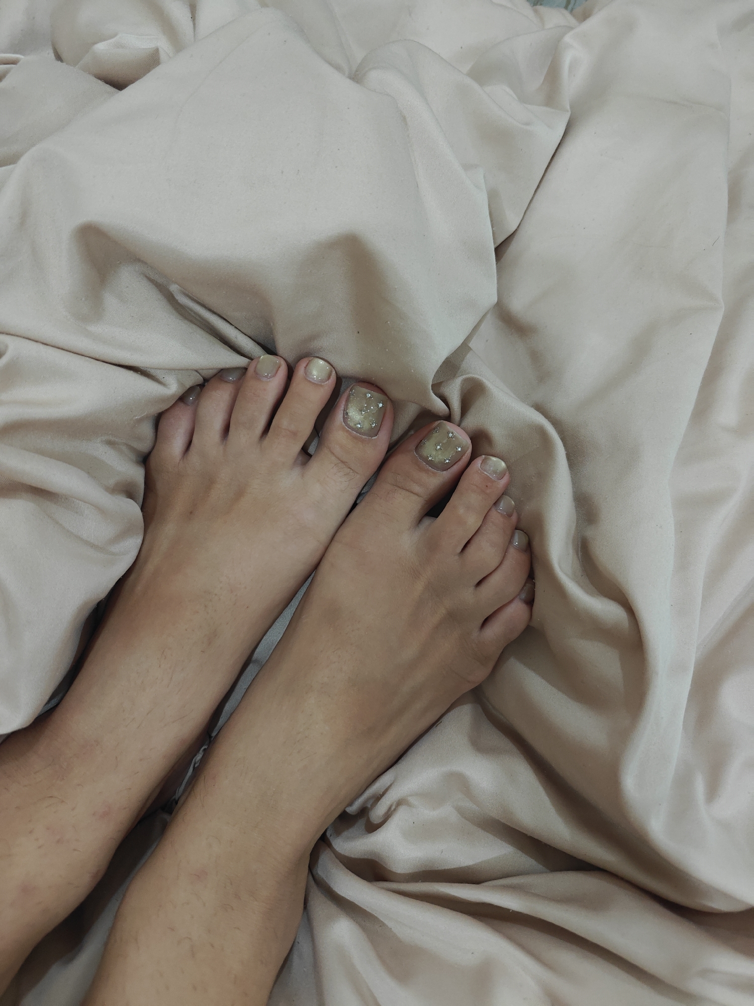 The image shows a close-up view of two feet, resting on a creased, light beige satin-like fabric, likely bedding. The feet have light-colored toenails, with the big toes on both feet decorated with glitter and small embellishments. The skin tone of the feet is light brown, and there is visible hair on the legs and feet. The fabric creates soft folds and shadows, suggesting a relaxed or indoor setting. The lighting appears to be soft and diffused, possibly from an indoor light source or natural light filtered through a window. There is no visible text or specific time of day or weather cues, but the intimate framing and focus on the feet suggest a personal or candid moment. The scene is set in Tay Nha Trang Ward, Vietnam.