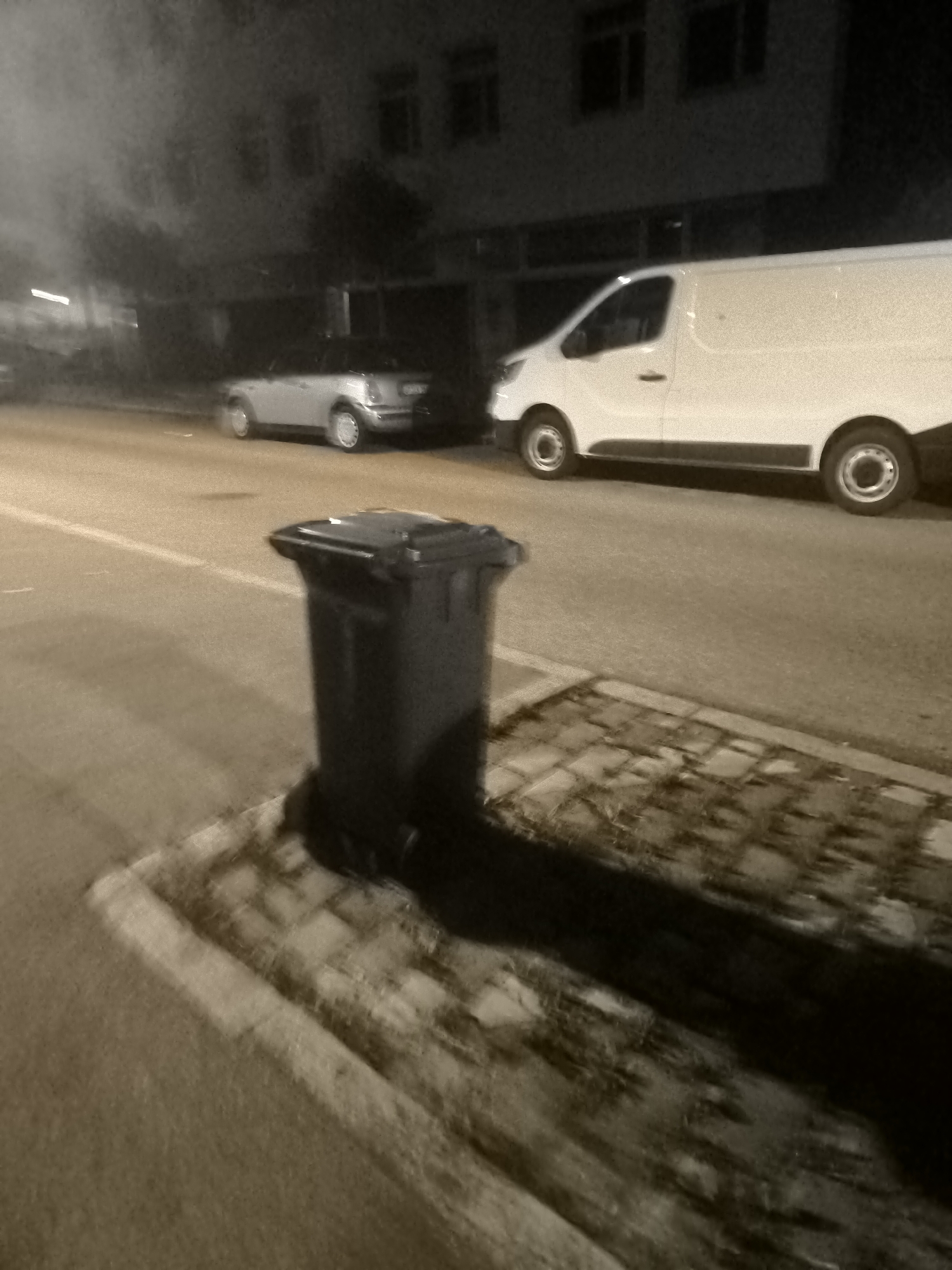 An outdoor, slightly blurry, nighttime photograph captures a street scene with a dark gray trash bin in the foreground. The bin is positioned on a paved area bordered by a curb and a sidewalk with cobblestone accents. In the mid-ground, a small silver car is parked on the left side of the road. Further down the street, a large white van is parked. The background features the facade of a building with dark windows. The overall lighting is dim, suggesting artificial illumination from streetlights or nearby buildings.